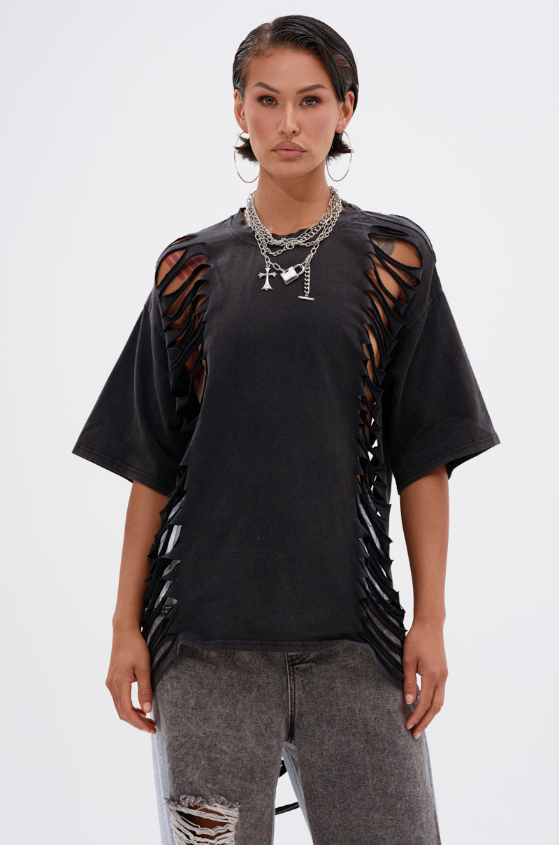 Model wears the SHAINA DISTRESSED OVERSIZED TEE with wide vertical cutouts on the sides and sleeves, paired with layered necklaces and high-waisted faded gray jeans, standing against a plain white background. 1