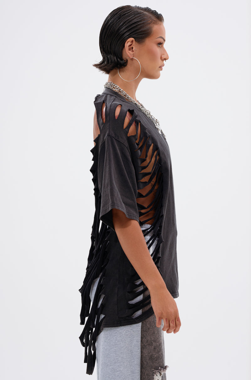 A person with short, slicked-back hair wears the SHAINA DISTRESSED OVERSIZED TEE, featuring dramatic side and back slashes, paired with hoop earrings and layered necklaces, standing in profile against a plain background. 2