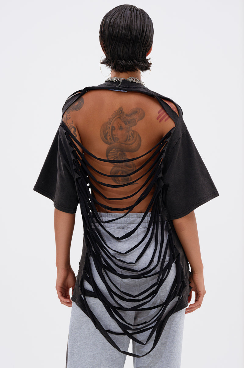 A person with wet, slicked-back hair wears the SHAINA DISTRESSED OVERSIZED TEE featuring a slashed, open back that reveals tattoos, paired with gray pants. The background is plain white. 3