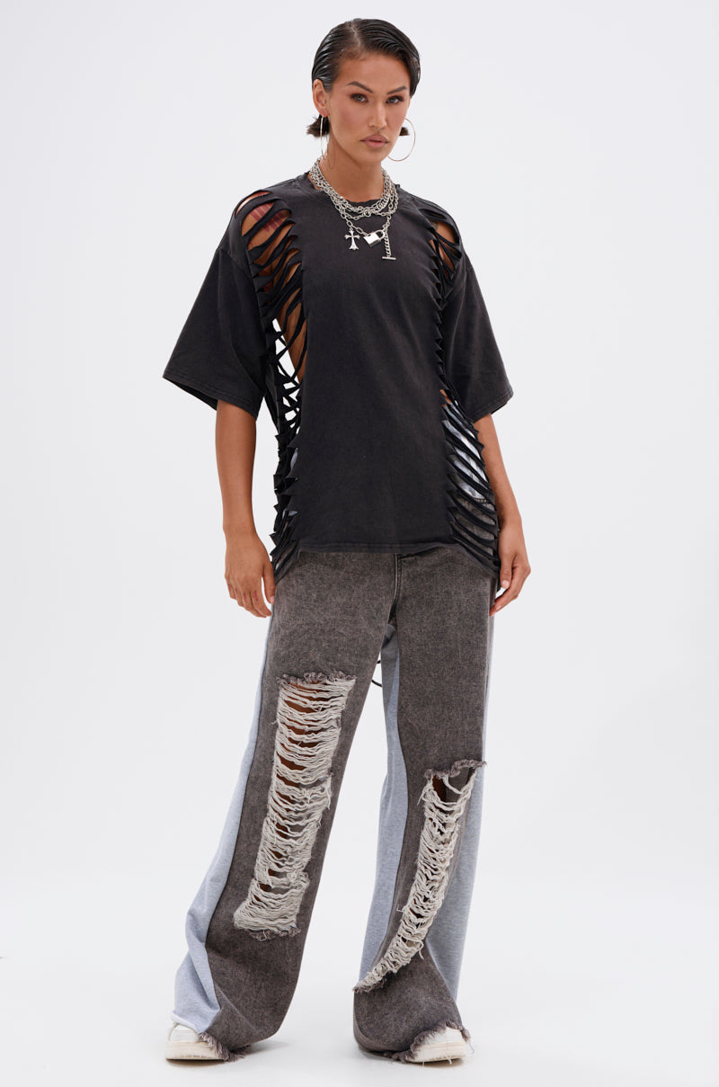 A person stands confidently in the SHAINA DISTRESSED OVERSIZED TEE, a black punk-inspired shirt with bold side and sleeve cutouts, paired with layered necklaces and loose wide-leg jeans featuring heavy front distressing. 4