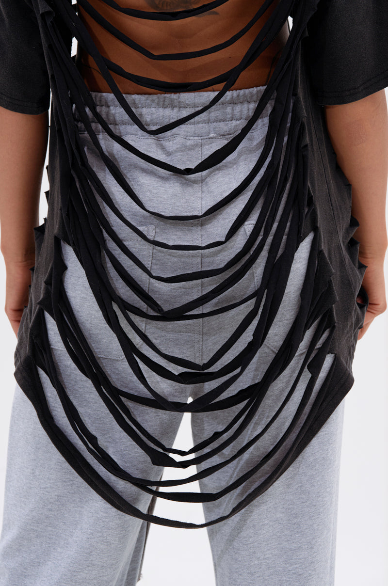 A person wears light gray sweatpants and the SHAINA DISTRESSED OVERSIZED TEE, a black, punk-inspired shirt with multiple horizontal cut-out strips on the back that reveal skin. The image is cropped from mid-back to thigh. 6
