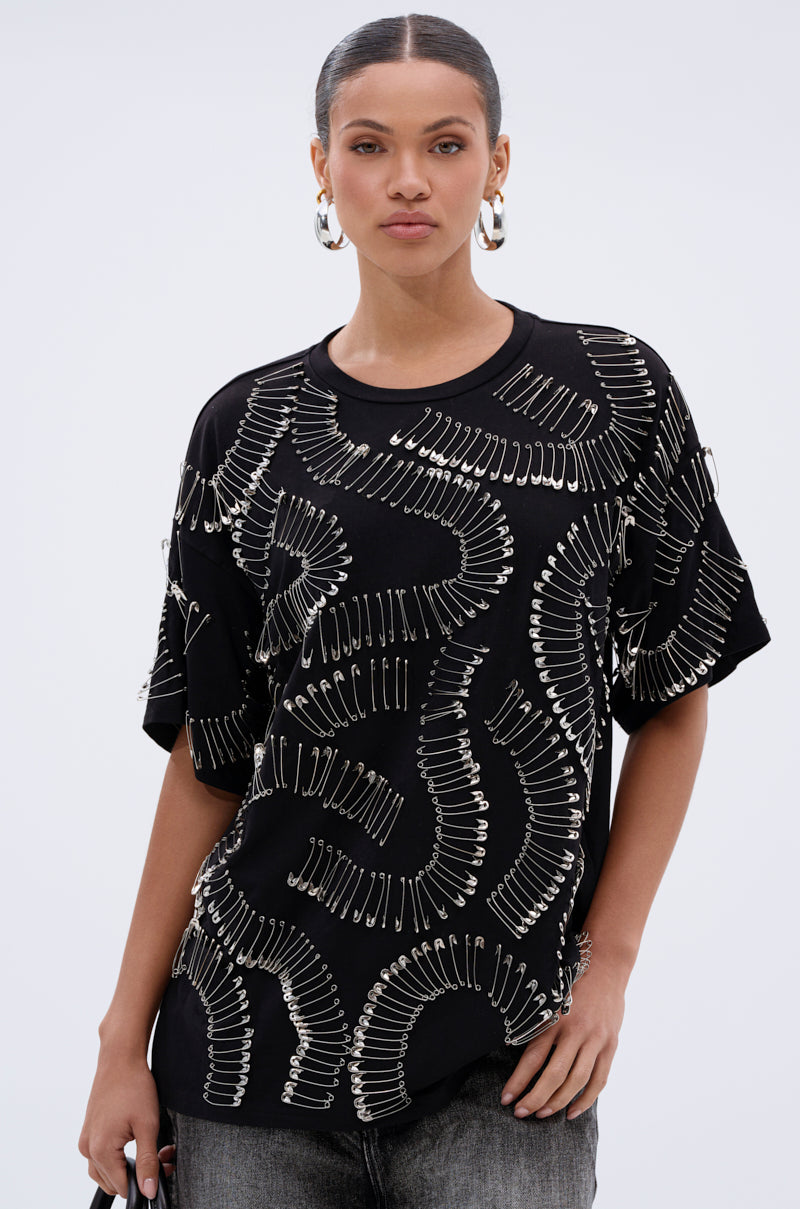A woman with pulled-back hair and large hoop earrings wears the SAFE AND SOUND OVERSIZED TEE, a black top adorned with swirling silver safety pin patterns, giving her a bold streetwear look as she faces forward with a neutral expression.
