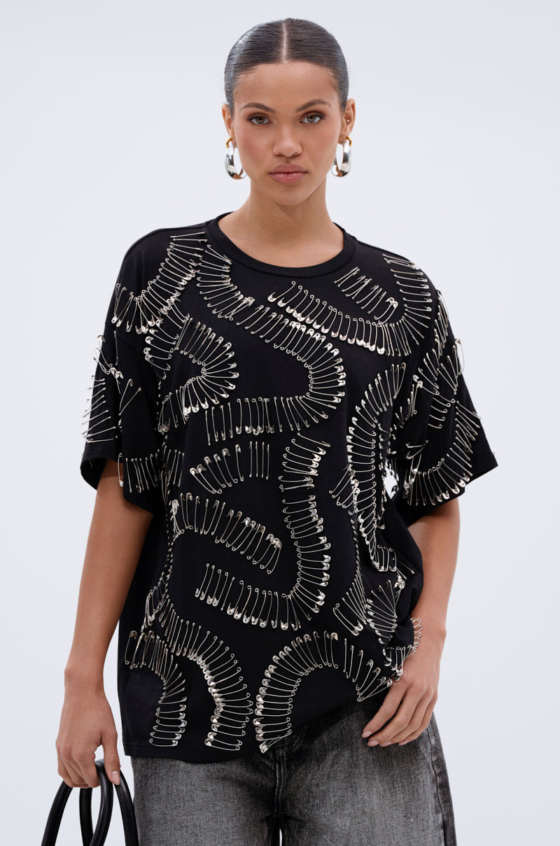 A woman with slicked-back hair wears the SAFE AND SOUND OVERSIZED TEE, featuring overlapping silver paper clip hardware, styled with gray jeans, a black bag, and large hoop earrings against a plain light background.