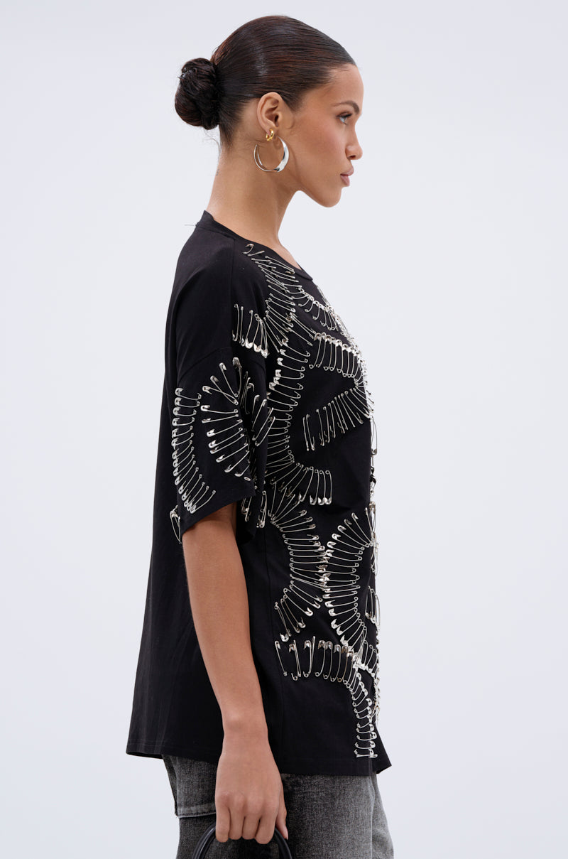 A woman in a low bun and large hoop earrings models the SAFE AND SOUND OVERSIZED TEE, featuring a white abstract pattern, standing in profile against a plain light gray background for a modern streetwear look.