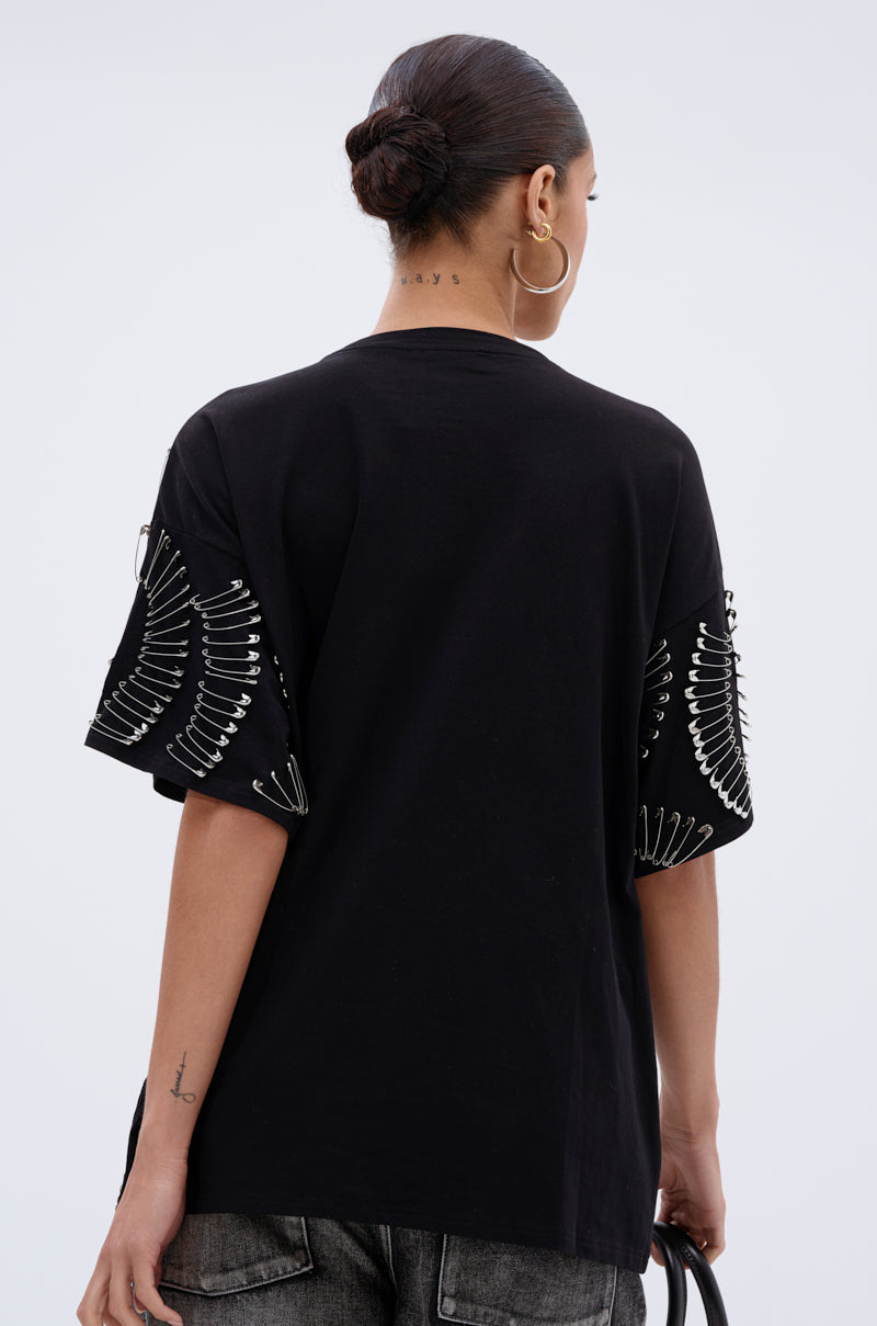 A person with hair in a low bun is shown from the back against a plain background, wearing the SAFE AND SOUND OVERSIZED TEE featuring silver paper clip hardware on the sleeves, dark jeans, hoop earrings, and holding a bag.