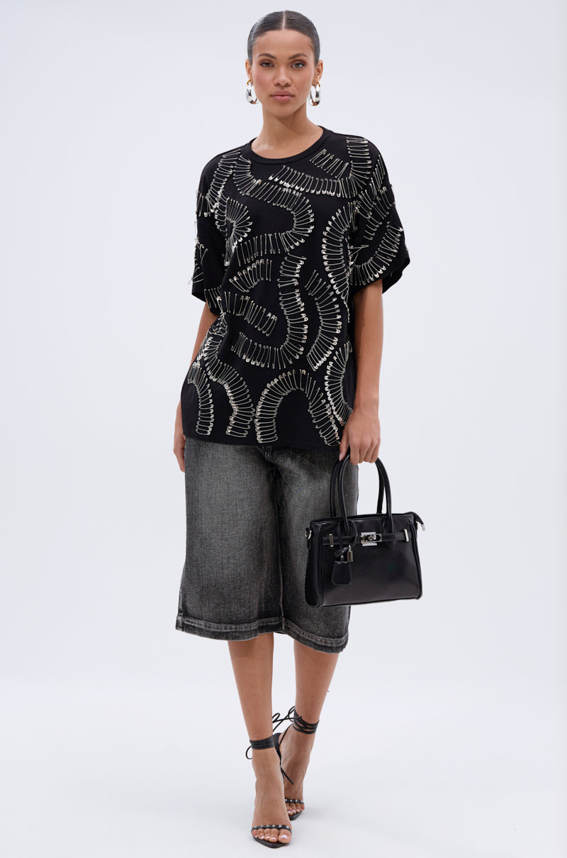 A woman wearing the SAFE AND SOUND OVERSIZED TEE, knee-length dark denim shorts, strappy heels, hoop earrings, and holding a black handbag stands against a plain white background.