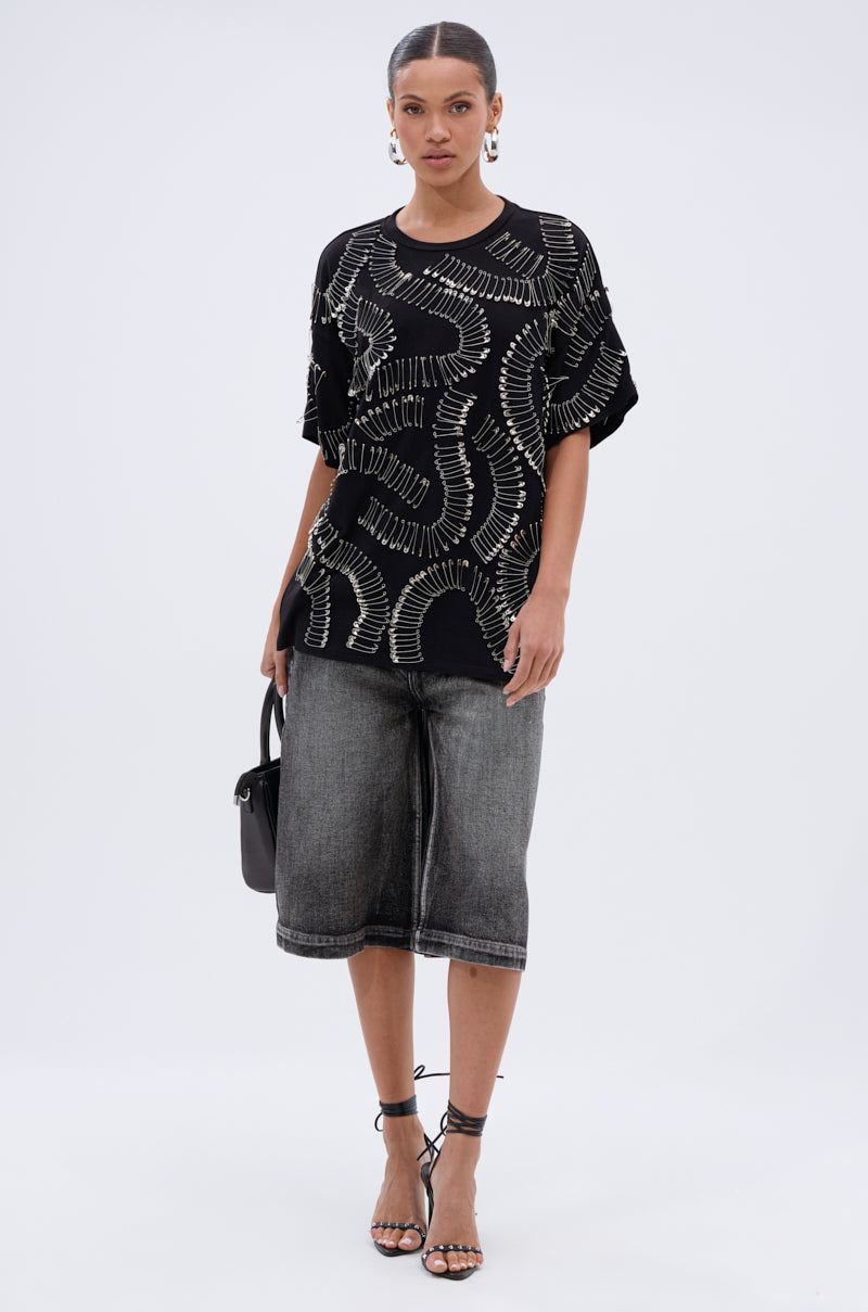 A woman stands against a plain background wearing the SAFE AND SOUND OVERSIZED TEE, loose faded black knee-length denim shorts, strappy black sandals, hoop earrings, and carrying a small black handbag.