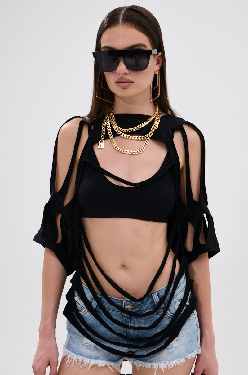 Wearing the NOTHING THERE DISTRESSED TEE, a woman in large black sunglasses, layered gold chains, and distressed denim shorts presents a deconstructed look with cut-out details against a plain white background.