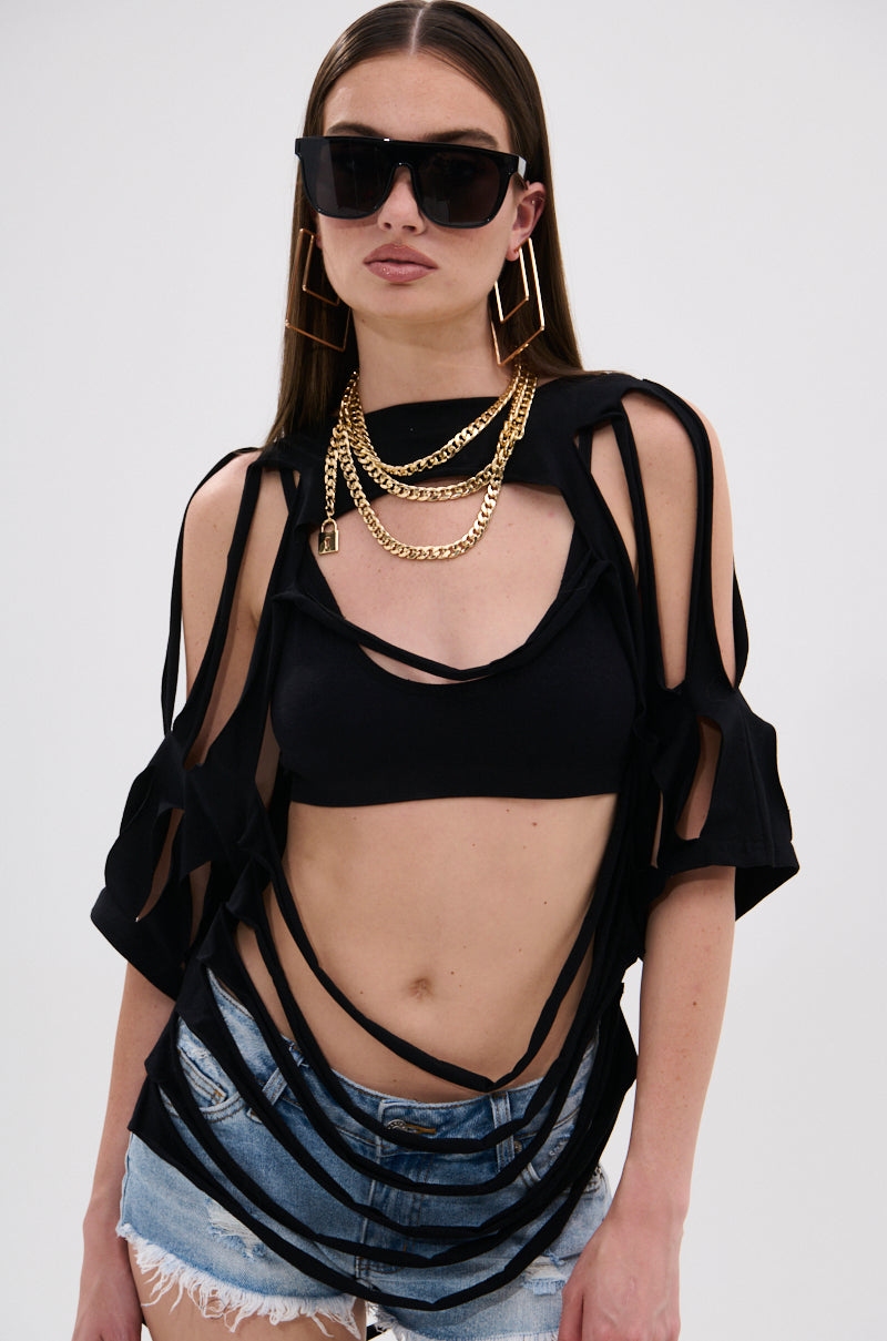 A woman models the NOTHING THERE DISTRESSED TEE over a black crop top, paired with black sunglasses, large gold earrings, layered gold necklaces, and distressed denim shorts against a plain backdrop.