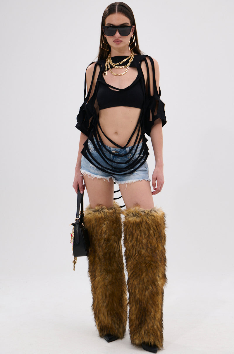 A woman stands against a white background wearing oversized sunglasses, gold jewelry, the NOTHING THERE DISTRESSED TEE, denim shorts, tall brown furry boots, and holding a small black handbag.