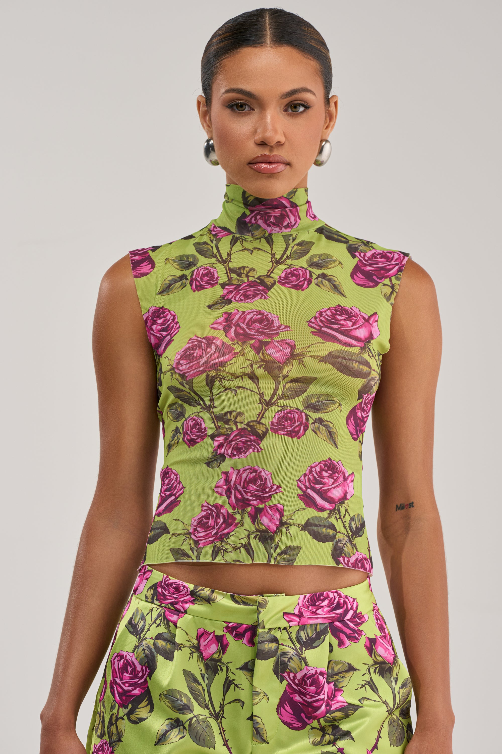 A woman stands against a plain background wearing the DOZEN ROSES MESH TOP IN CHARTREUSE, a sleeveless mock neck style in green with pink rose print. She pairs it with matching pants, hoop earrings, and has her hair slicked back.