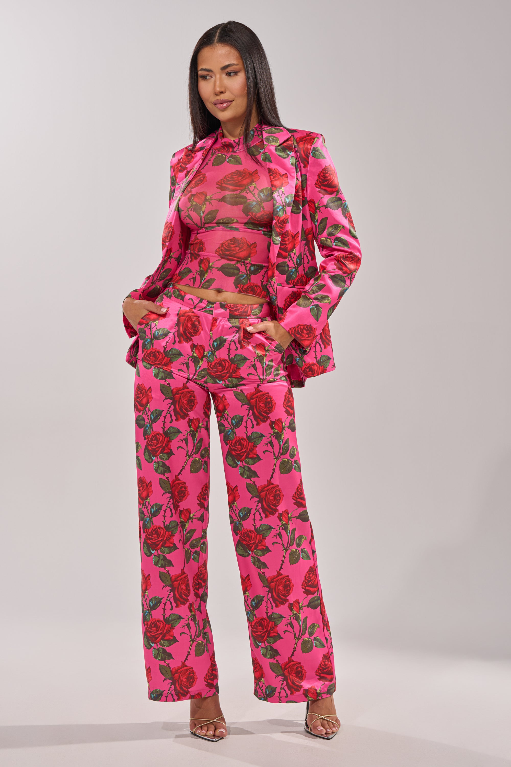 A woman poses in the DOZEN ROSES MESH TOP—bright pink with bold red rose print—styled with a matching blazer and wide-leg trousers, her straight hair down and hands in her pockets against a plain backdrop. 9