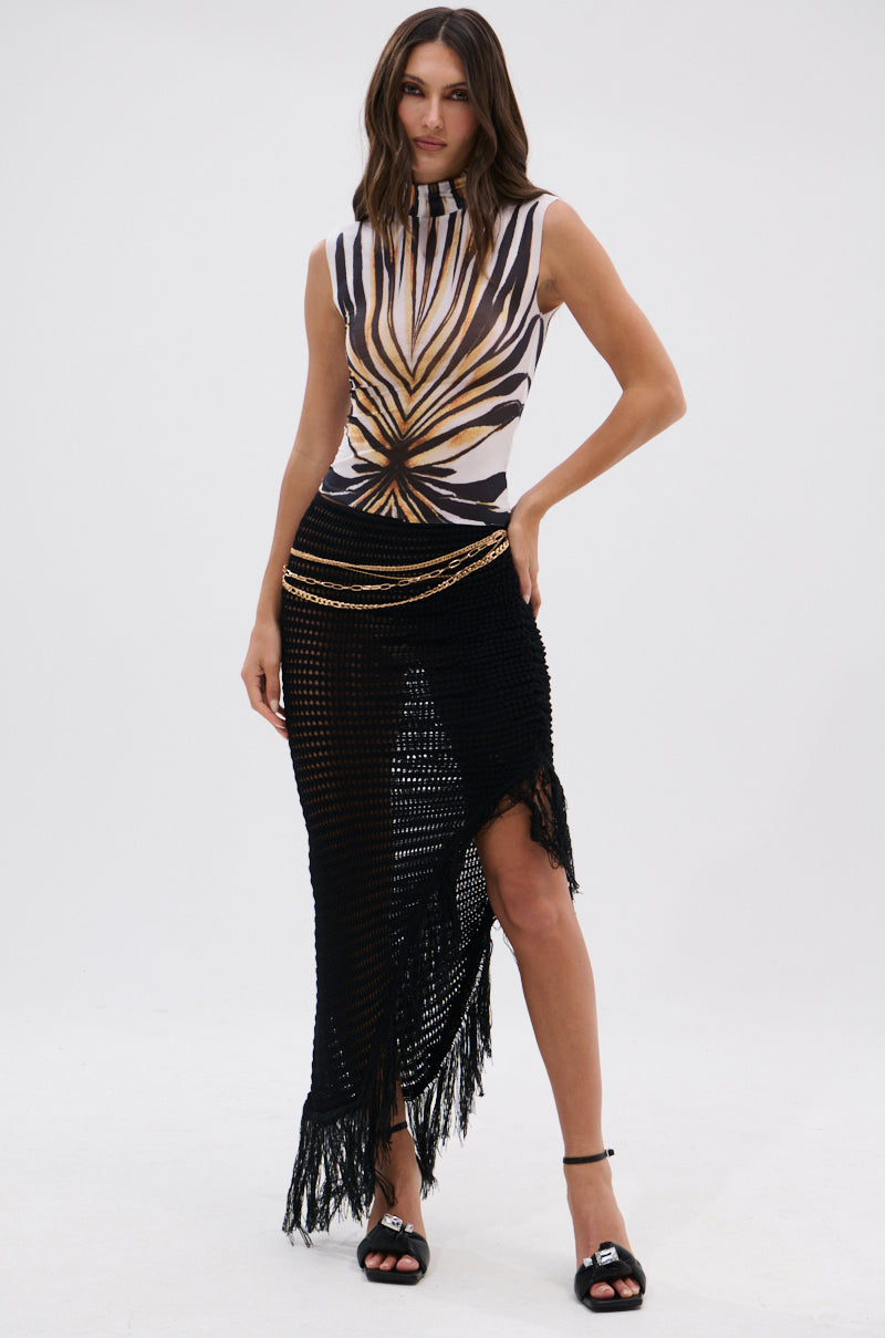 A woman models the PRIMAL SPIRIT SLEEVELESS MESH TOP with a bold zebra print, paired with a black asymmetric crochet skirt featuring fringe detail and black heeled sandals, standing against a plain white background. 7