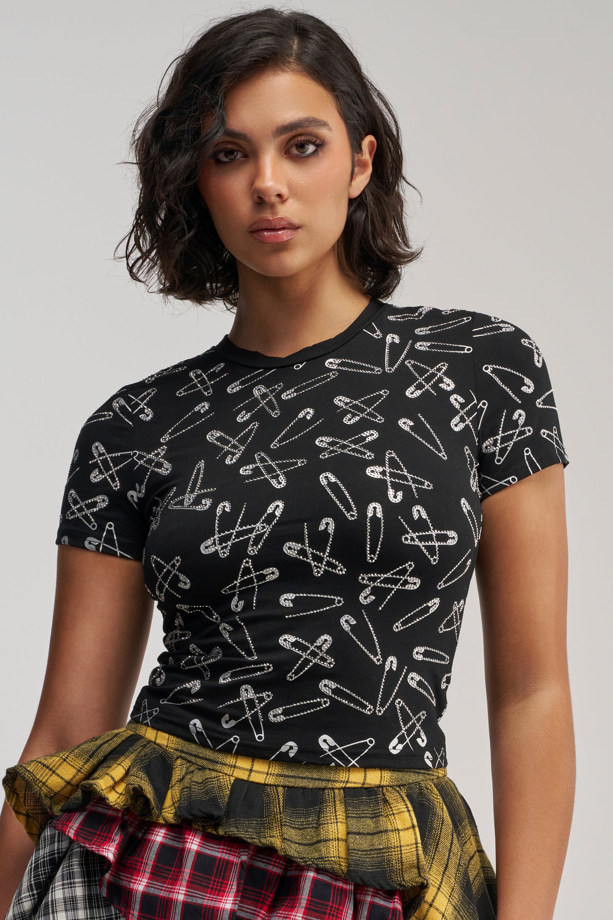 A woman with short dark hair wears the PROCEED WITH CAUTION RHINESTONE TEE, a streetwear-inspired black t-shirt adorned with white safety pin patterns, paired with a plaid skirt featuring yellow and red layers, as she stands against a plain background.
