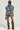 Wearing the OVERRATED PLAID MINI SKIRT featuring a ruffled design and denim trim waistband, along with sunglasses and tall brown slouch boots, the person holds a small brown purse while standing against a plain background.