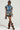 A woman wears the OVERRATED PLAID MINI SKIRT, paired with a blue and brown plaid ensemble that includes a denim crop top with a denim trim waistband. She accessorizes her outfit with a small handbag, oversized sunglasses, and knee-high brown boots featuring ruffles.