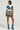 A fashionable person stands confidently against a light background, wearing a white shirt with a cropped denim vest and the OVERRATED PLAID MINI SKIRT featuring a denim trim waistband, complemented by blue denim boots and dark sunglasses.