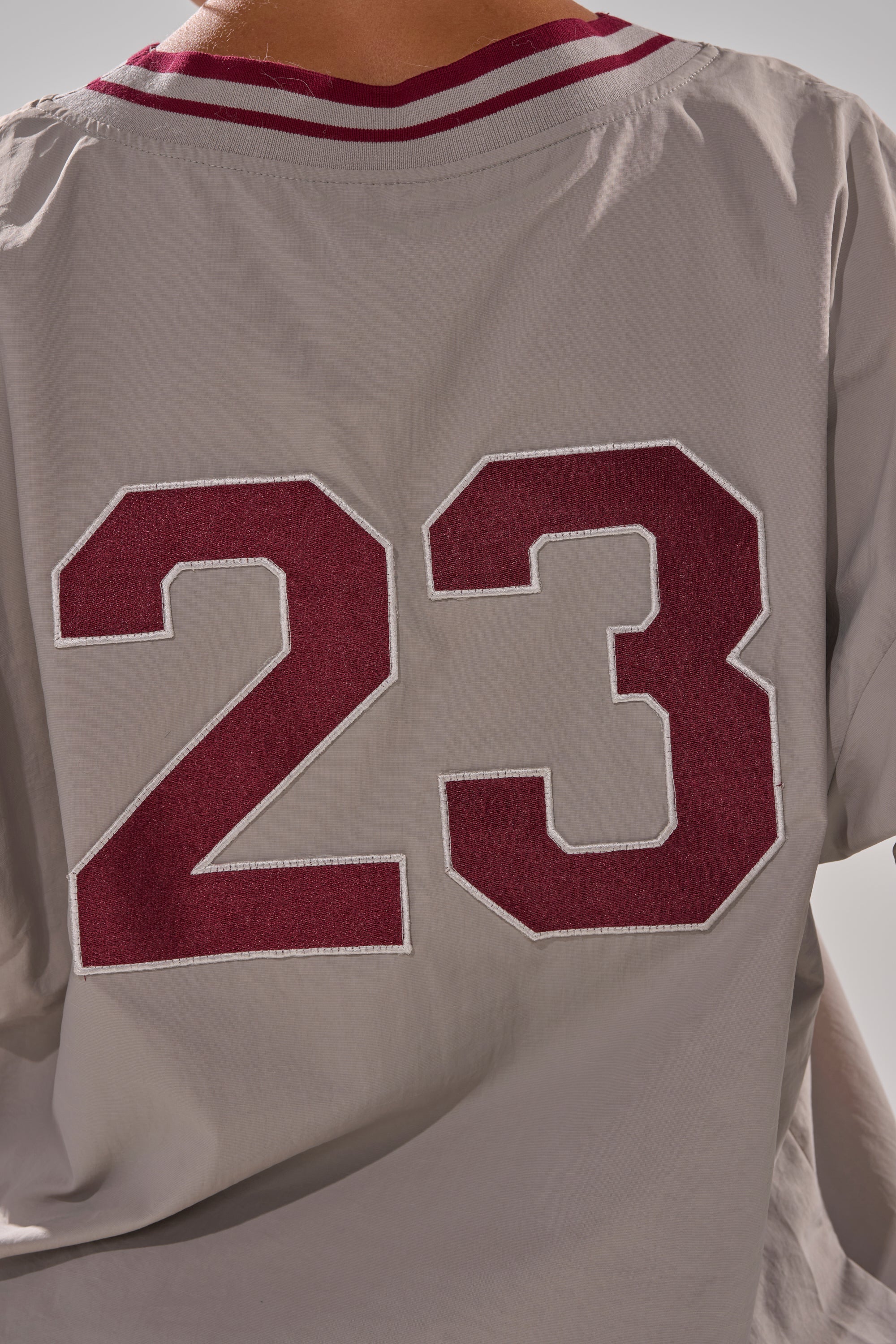 A person wears a light gray CHICAGO V-NECK SWEATSHIRT jersey featuring a large maroon and white outlined number 23 on the back, with a maroon and white striped collar and an oversized silhouette.