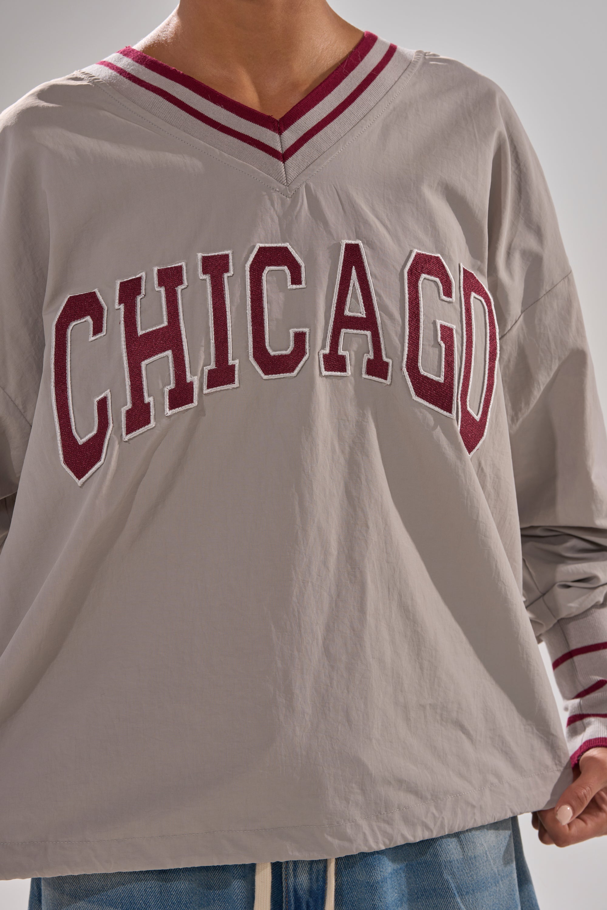 Someone wearing the CHICAGO V-NECK SWEATSHIRT—an oversized, light gray top with maroon stripes and bold "CHICAGO" lettering across the chest; only their torso is visible.