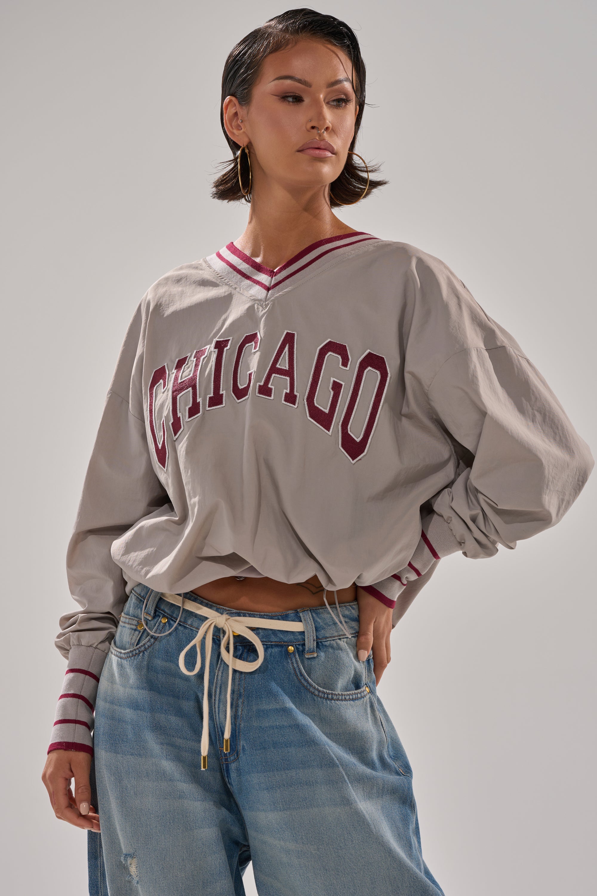 A person stands confidently against a plain background, wearing the CHICAGO V-NECK SWEATSHIRT in oversized gray windbreaker fabric, paired with blue jeans featuring a rope belt and large hoop earrings.