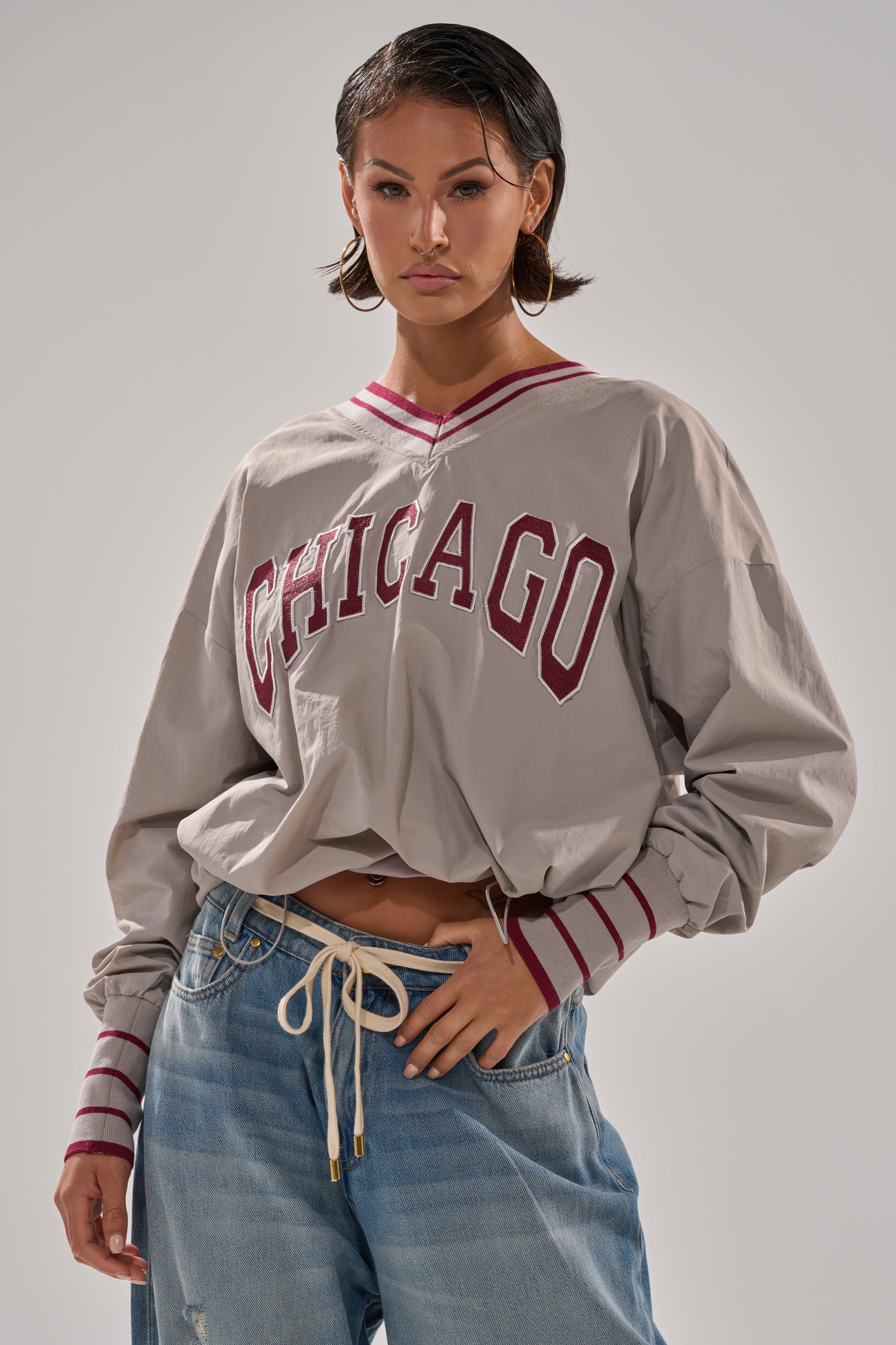 A person in hoop earrings and light blue jeans wears the gray CHICAGO V-NECK SWEATSHIRT with red lettering, standing against a plain background with one hand in their pocket.