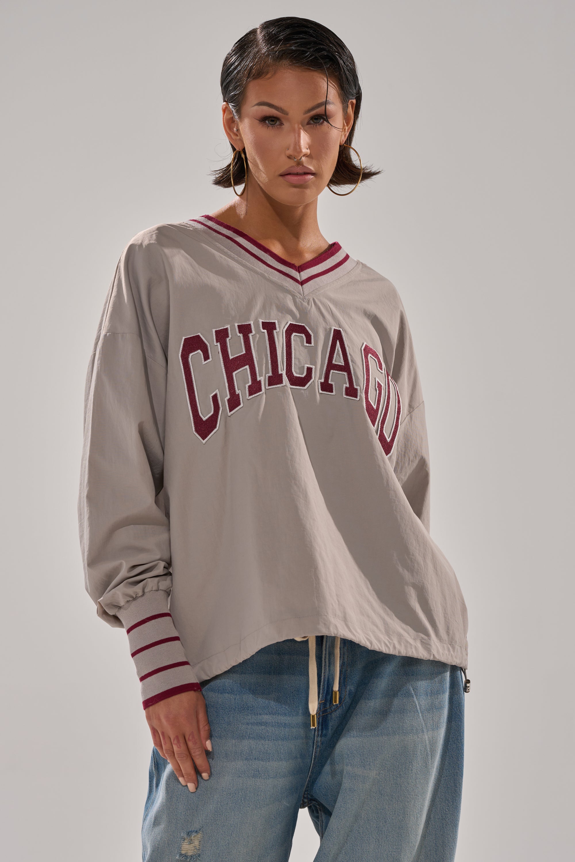 Someone sports the CHICAGO V-NECK SWEATSHIRT—loose, beige, with red-striped cuffs and bold red "CHICAGO" lettering—paired with blue jeans, short hair, and hoop earrings for a stylish ensemble.