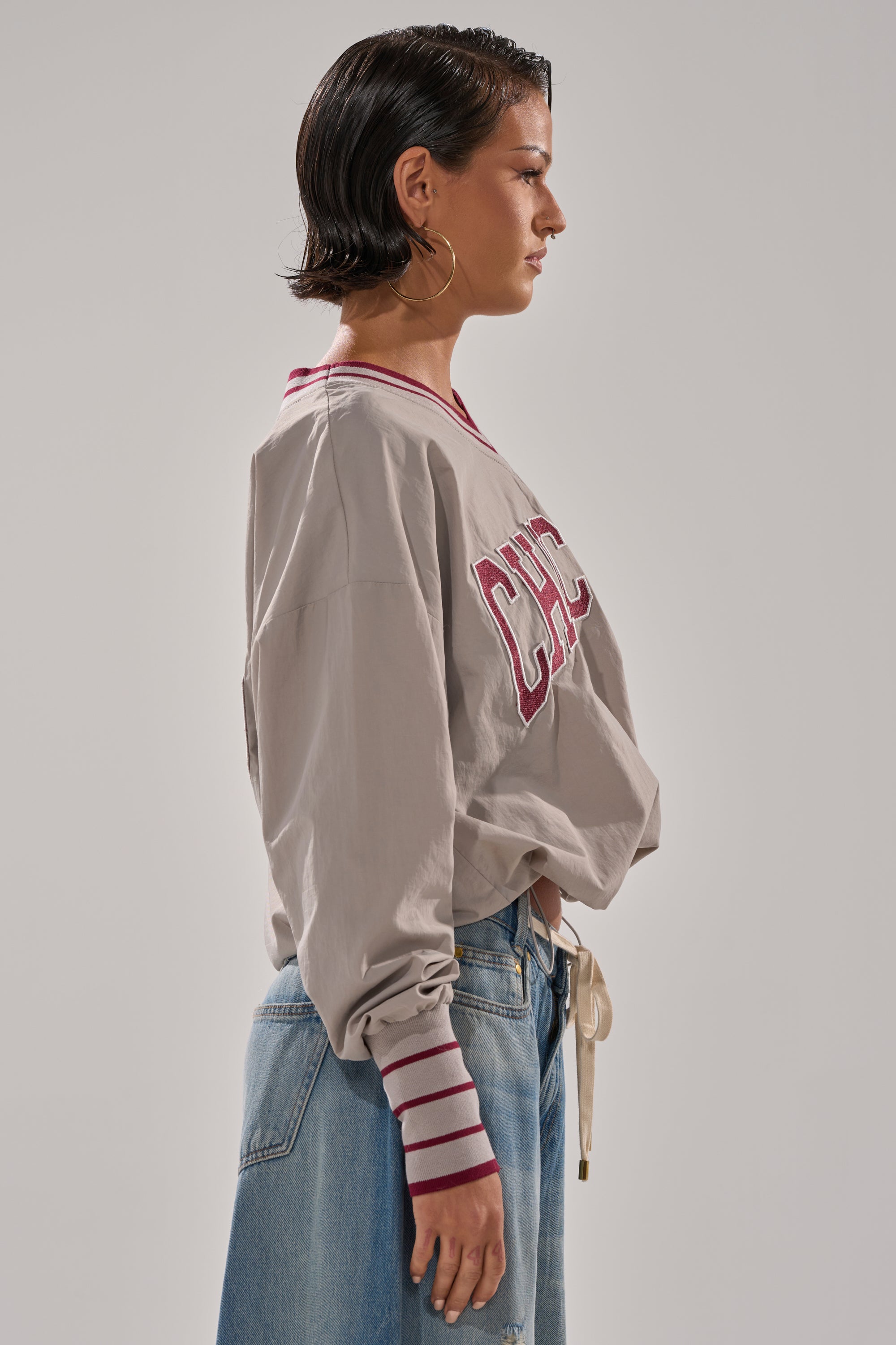 A person with short dark hair models the CHICAGO V-NECK SWEATSHIRT, featuring red lettering and striped cuffs, paired with loose blue jeans. They stand sideways against a plain gray background.