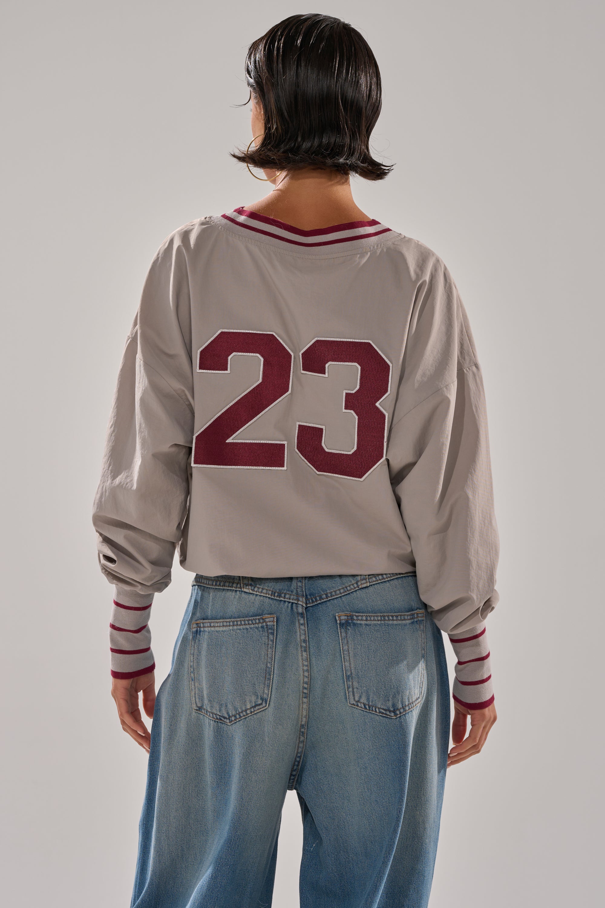 A person with short dark hair, wearing a CHICAGO V-NECK SWEATSHIRT featuring red stripes and the number 23 on the back, along with loose blue jeans, stands facing away from the camera.
