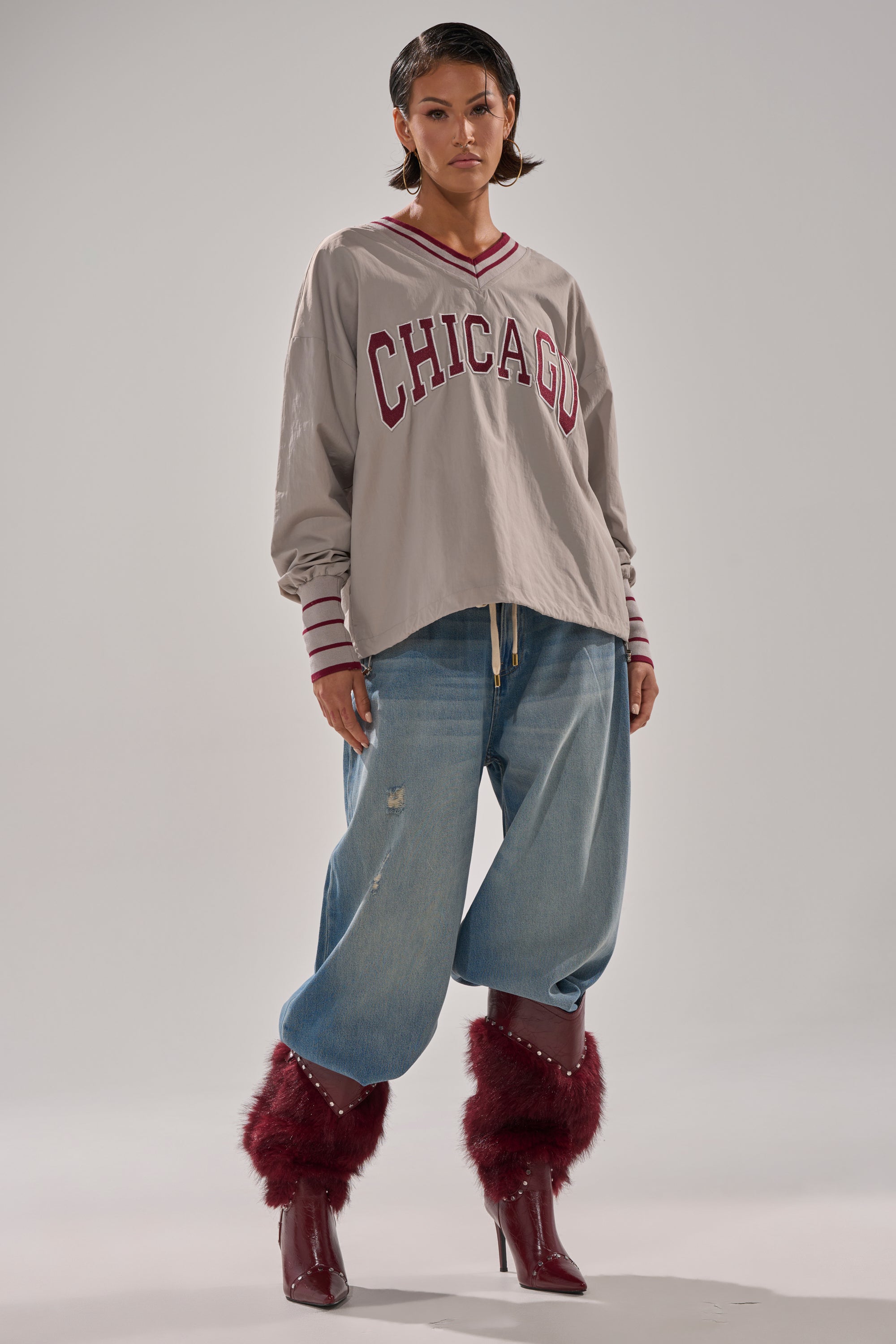 A person stands confidently in an oversized beige CHICAGO V-NECK SWEATSHIRT, paired with baggy ripped jeans and maroon fur-trimmed pointed-heel boots, against a plain light background.