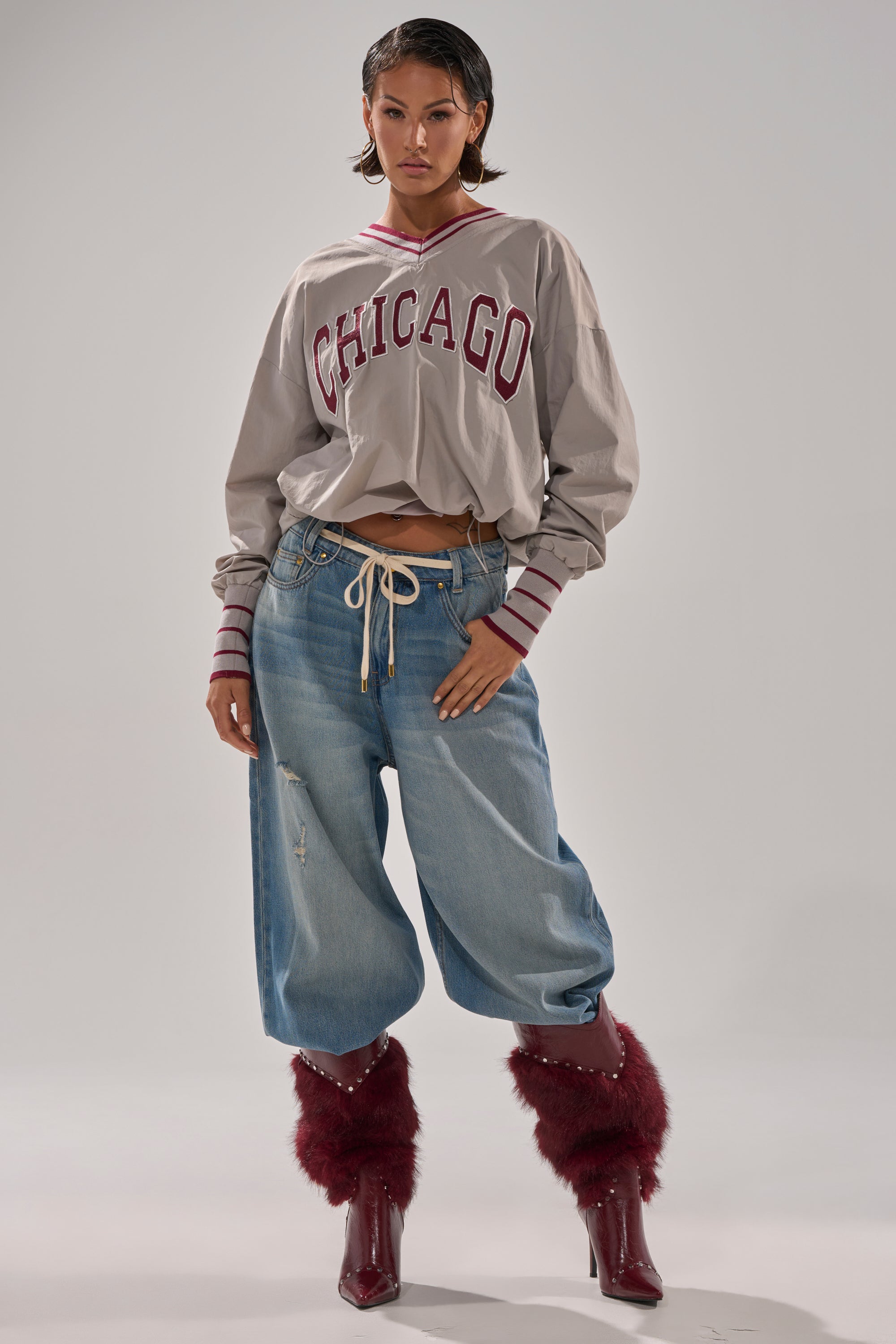 A woman poses confidently in the oversized CHICAGO V-NECK SWEATSHIRT, styled with loose ripped jeans and maroon furry pointed-heel boots, set against a plain grey background.