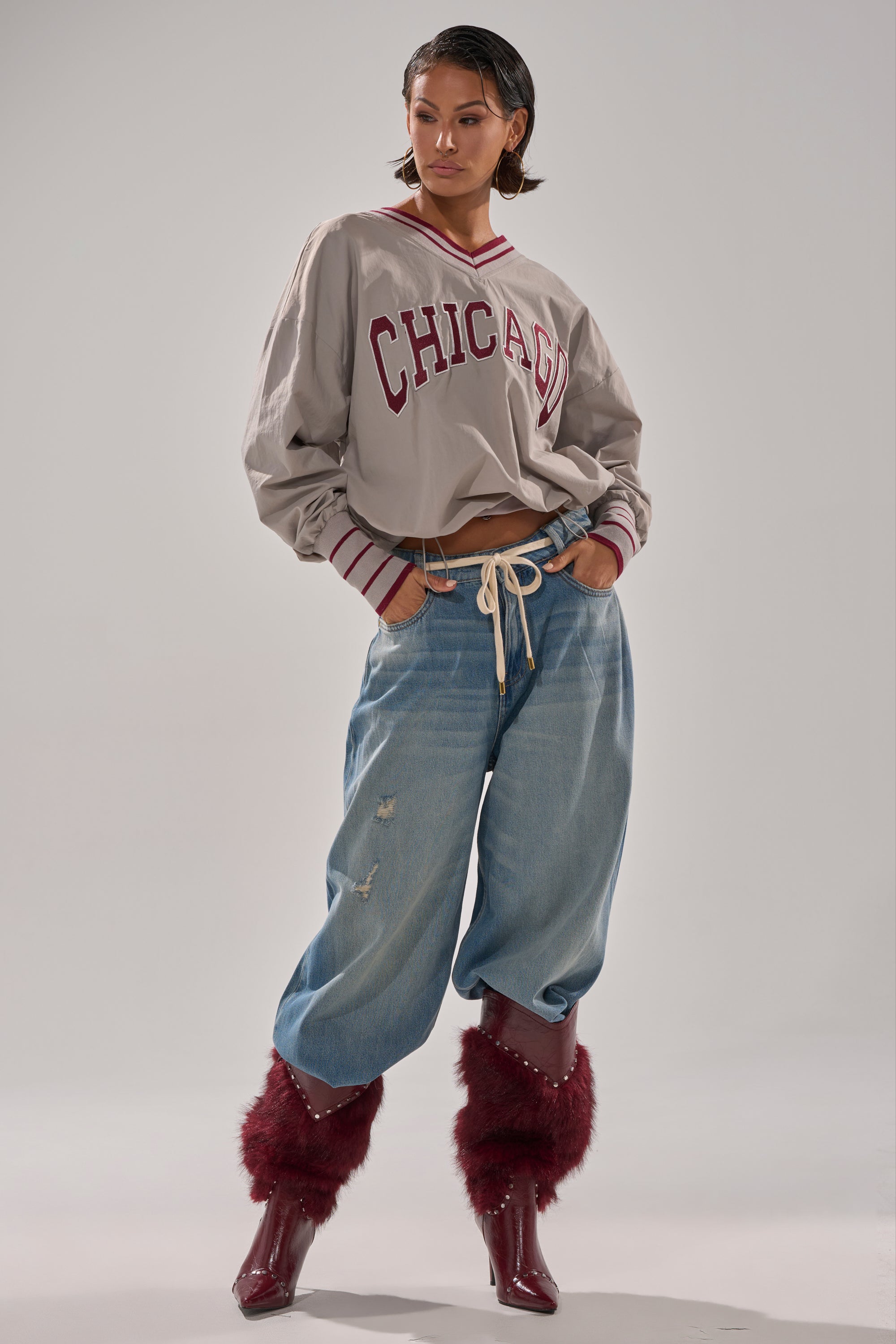 A person poses confidently against a gray background, wearing an oversized CHICAGO V-NECK SWEATSHIRT, loose ripped jeans with a rope belt, and shiny maroon boots with fluffy trim.