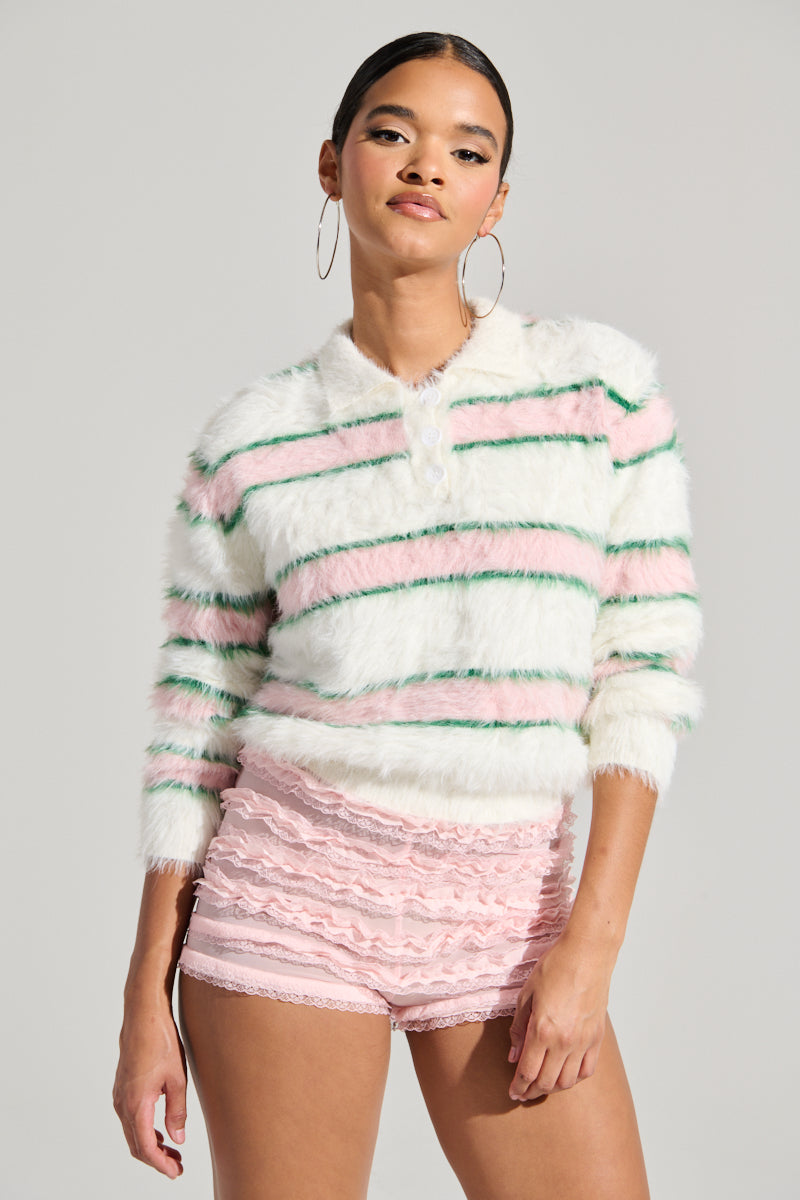A person wears the POLO PRINCESS STRIPED CROPPED SWEATER featuring green, pink, and white stripes, paired with pink lace shorts. They confidently pose against a plain gray background, accessorized with large hoop earrings.