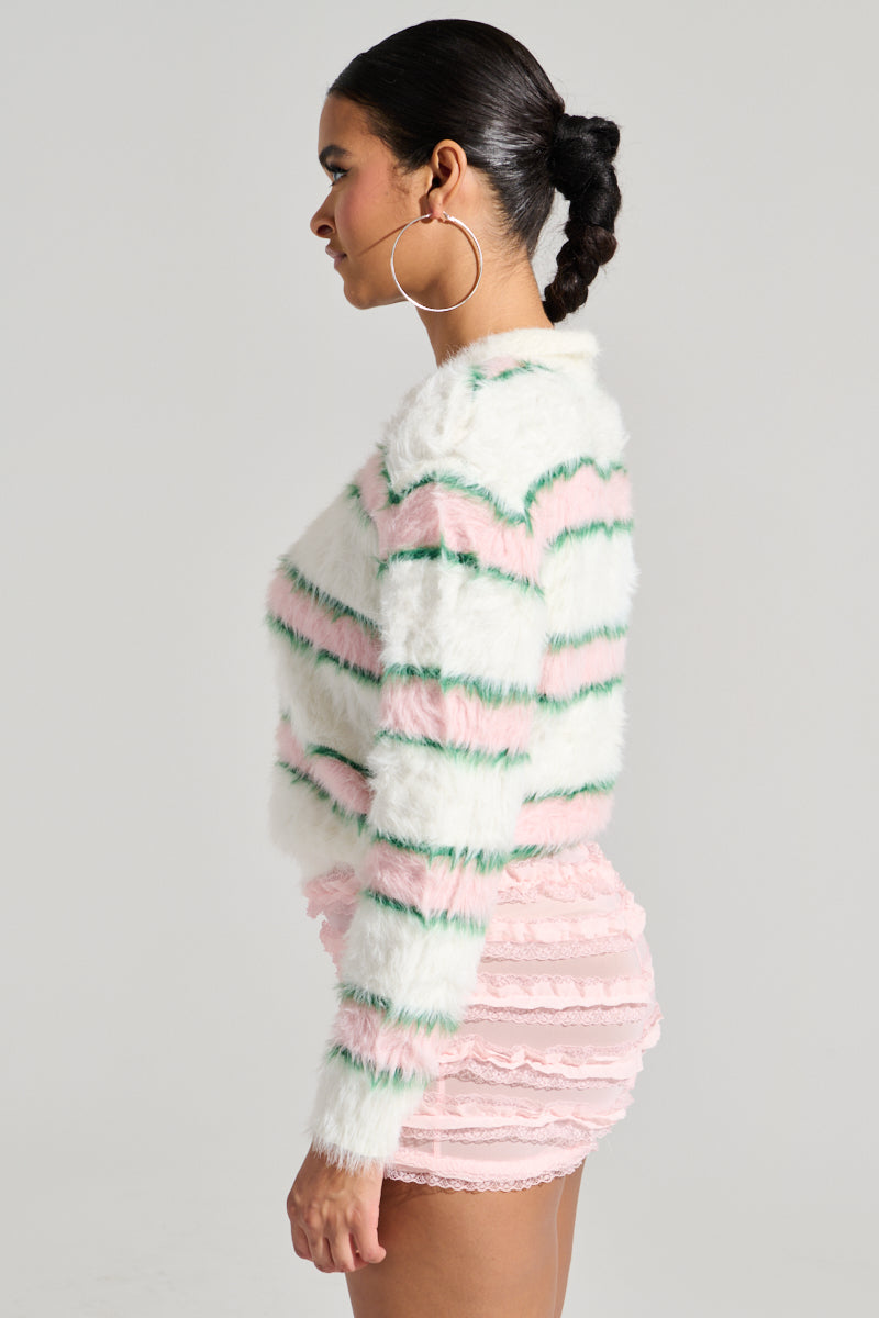 A woman is pictured in side profile wearing the POLO PRINCESS STRIPED CROPPED SWEATER, characterized by its fluffy texture and pink, green, and white stripes. She pairs the sweater with a pink lace skirt and accessorizes her look with large hoop earrings. Her hair is elegantly styled in a braided bun against a plain gray background.