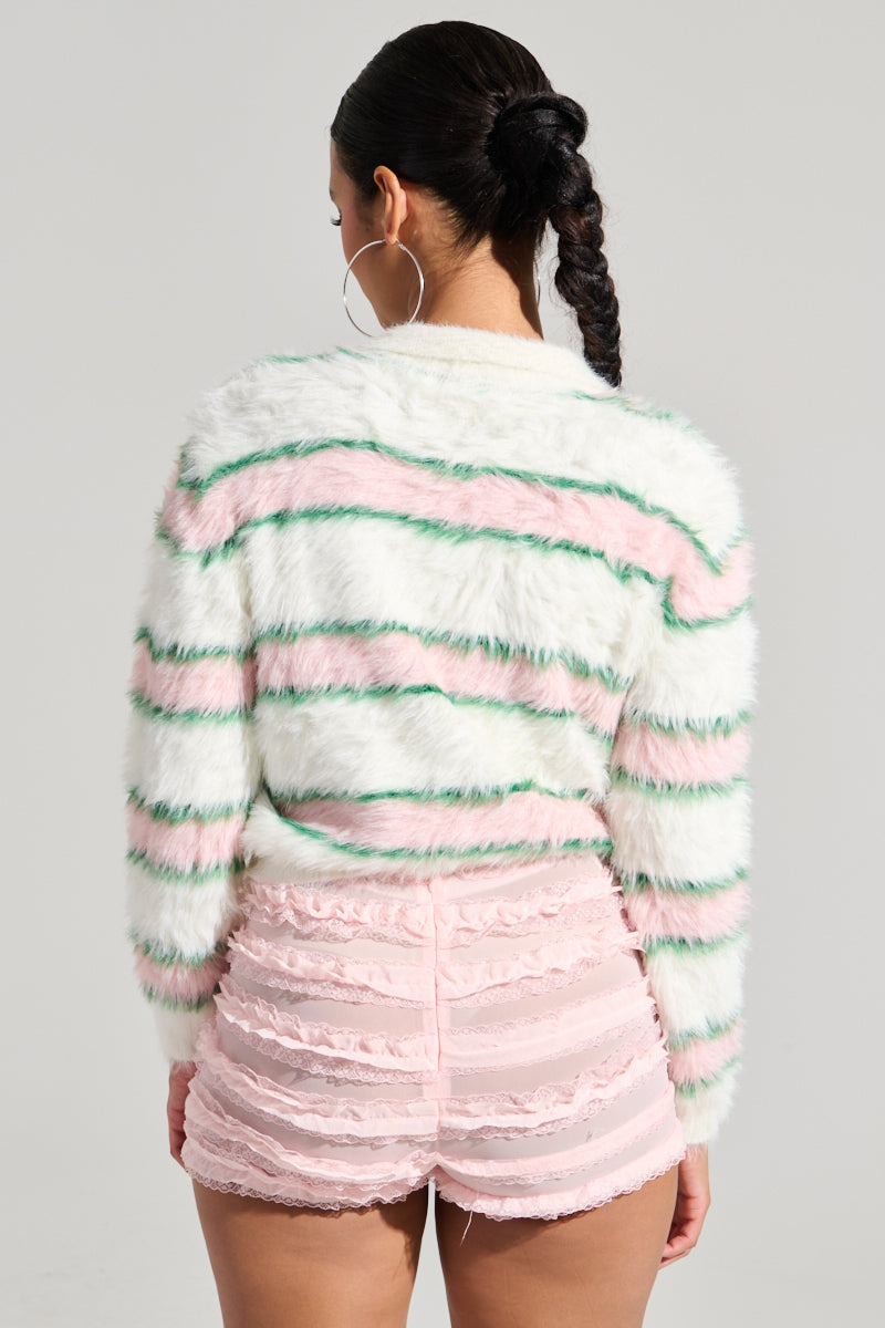 A person with a braided hairstyle is wearing the POLO PRINCESS STRIPED CROPPED SWEATER, which showcases pink and green stripes, paired with ruffled pink shorts. Large hoop earrings complete the look, set against a plain and neutral background.