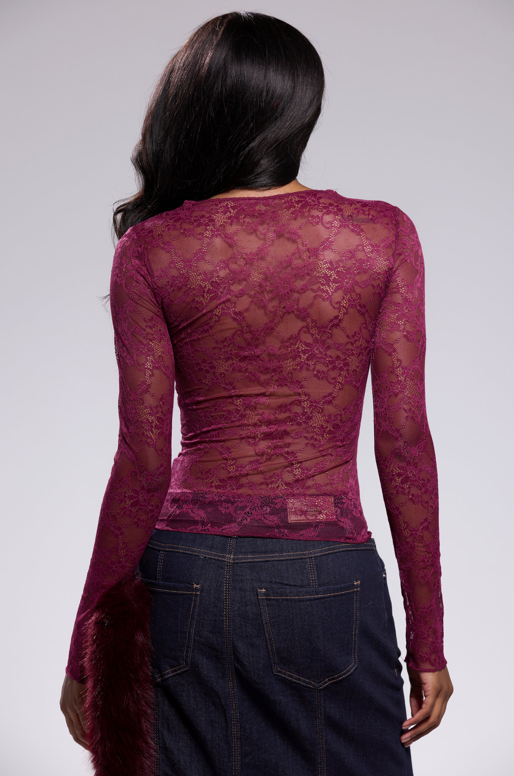 A woman with long dark hair, seen from the back, wears the ESSENTIAL LACE LONG SLEEVE TOP IN PLUM over a dark denim skirt against a plain light gray background. 3