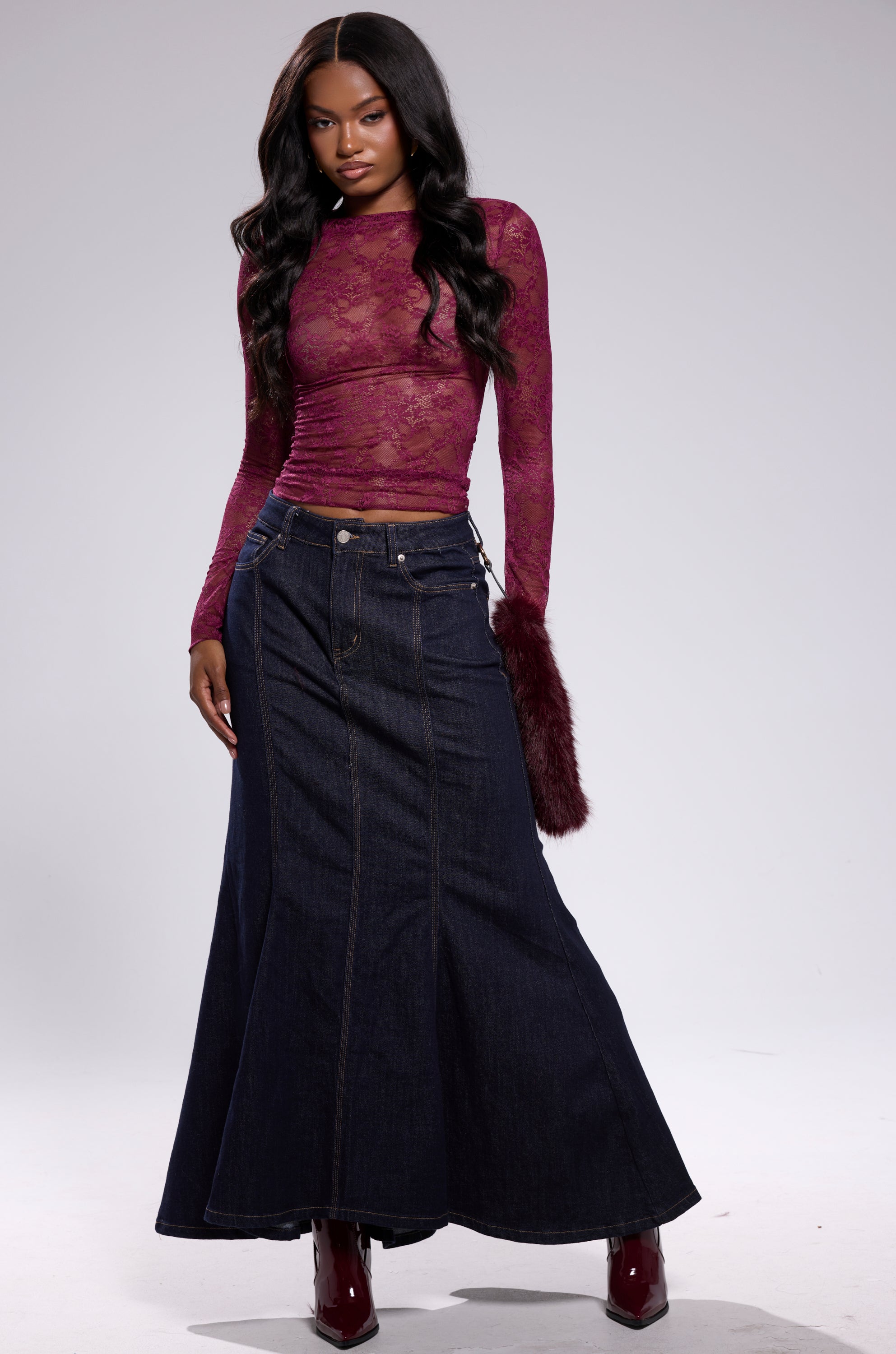 A woman stands confidently in the ESSENTIAL LACE LONG SLEEVE TOP IN PLUM, paired with a dark denim maxi skirt and burgundy boots. She holds a furry maroon accessory, creating a chic fall look against a plain light gray background. 5