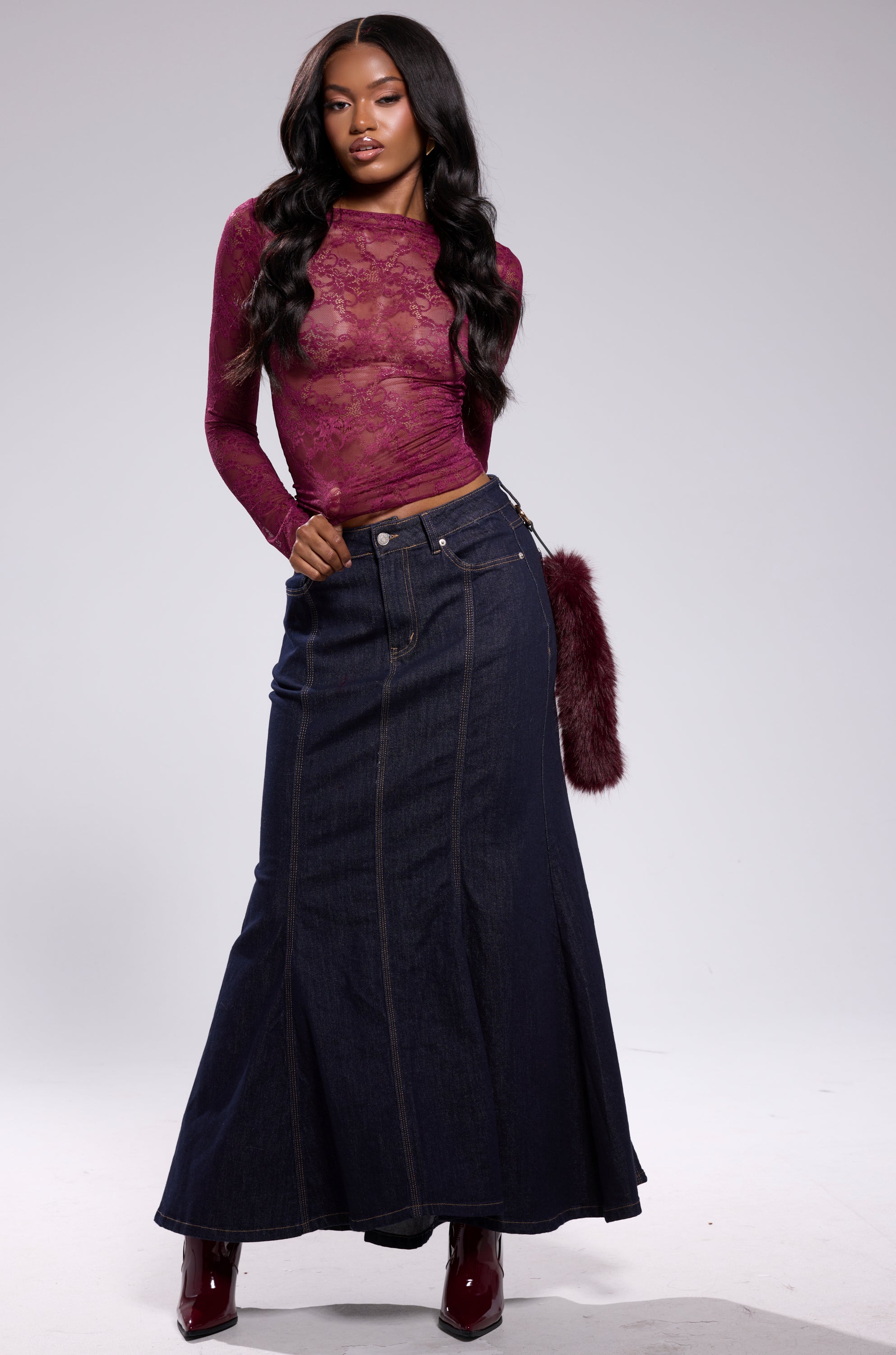 A woman poses in the ESSENTIAL LACE LONG SLEEVE TOP IN PLUM, paired with a high-waisted dark blue denim maxi skirt and burgundy boots. She has long wavy hair and stands against a plain light background. 6