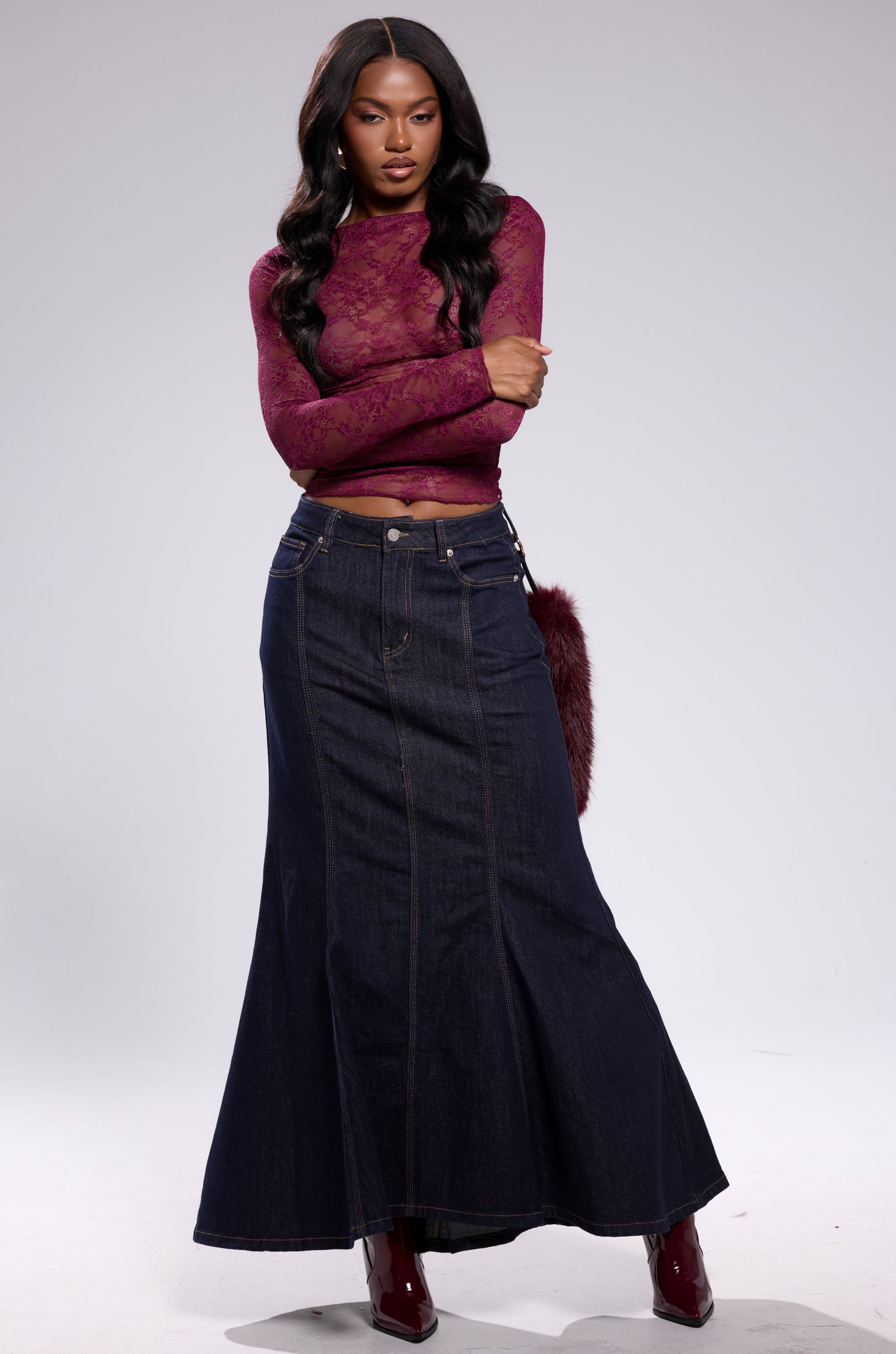 A woman with long, wavy hair stands against a plain background, arms crossed and looking at the camera. She wears the ESSENTIAL LACE LONG SLEEVE TOP IN PLUM with a high-waisted dark blue denim maxi skirt and maroon boots. 7