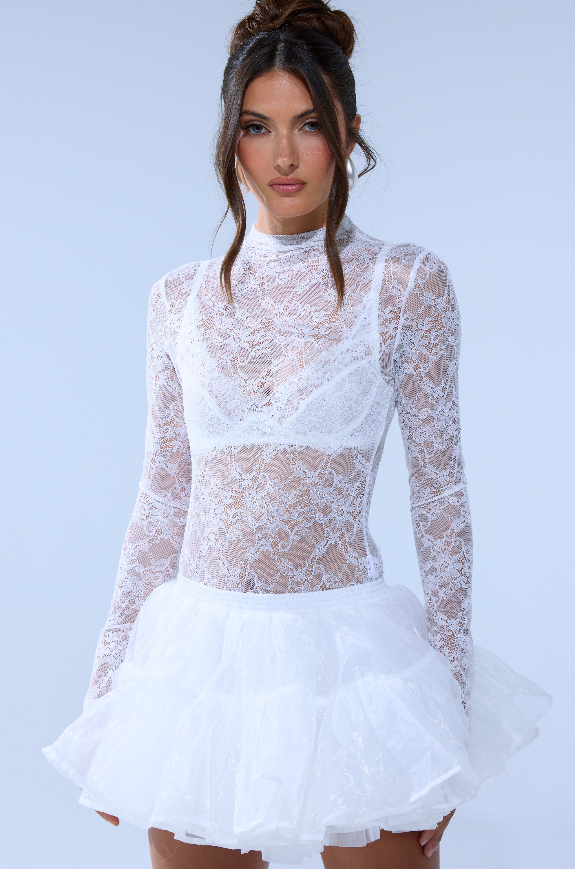 A woman models the EVERYDAY MOCK NECK LACE BODYSUIT IN WHITE with long sleeves over a white bra, paired with a tulle skirt. Her hair is styled in two buns with loose waves as she poses against a plain, light background. 1