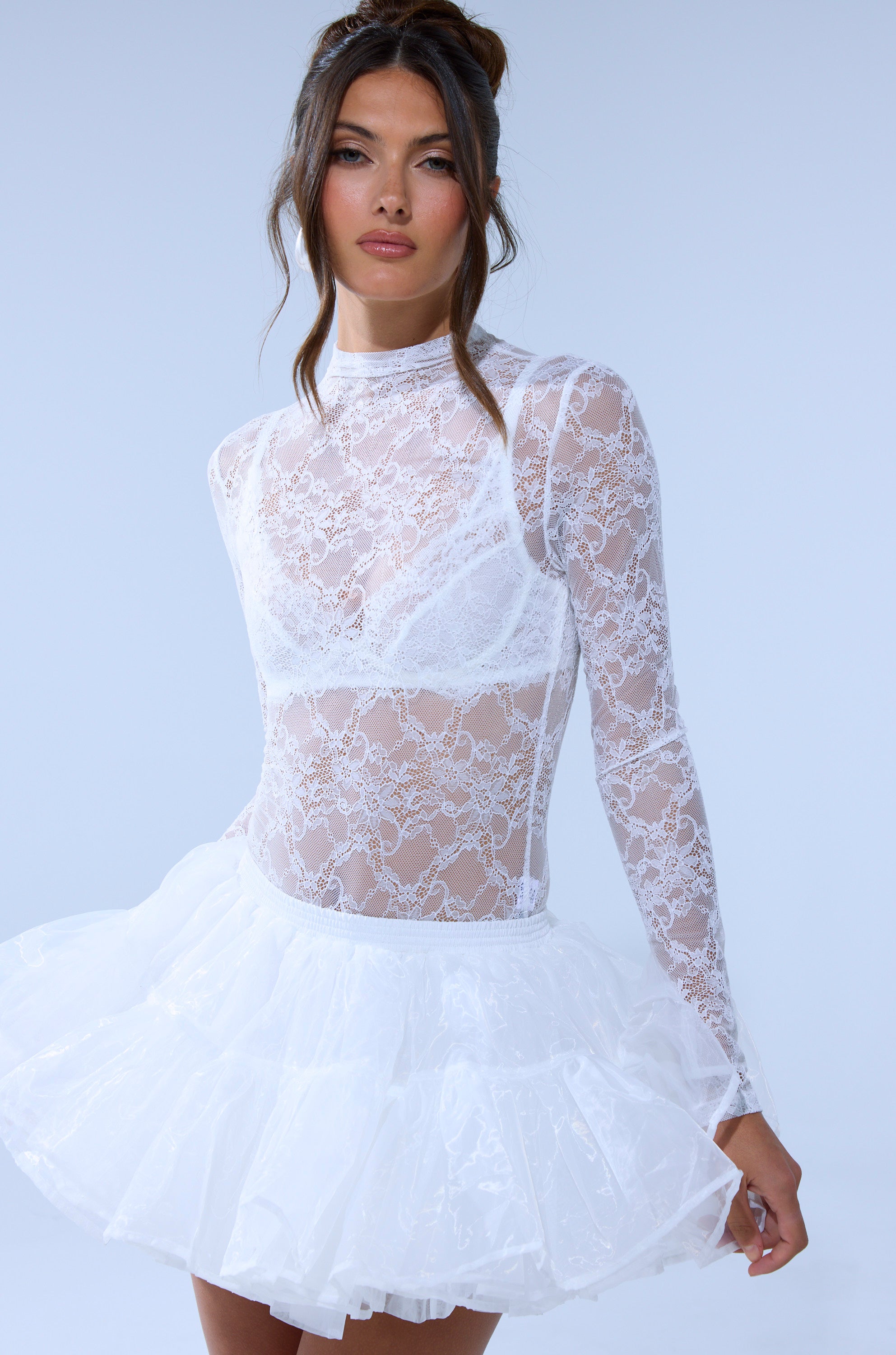 A woman wears the EVERYDAY MOCK NECK LACE BODYSUIT IN WHITE with sheer long sleeves over a white bra, styled with a short puffy tulle skirt. She stands against a plain light blue background, looking confidently at the camera. 2