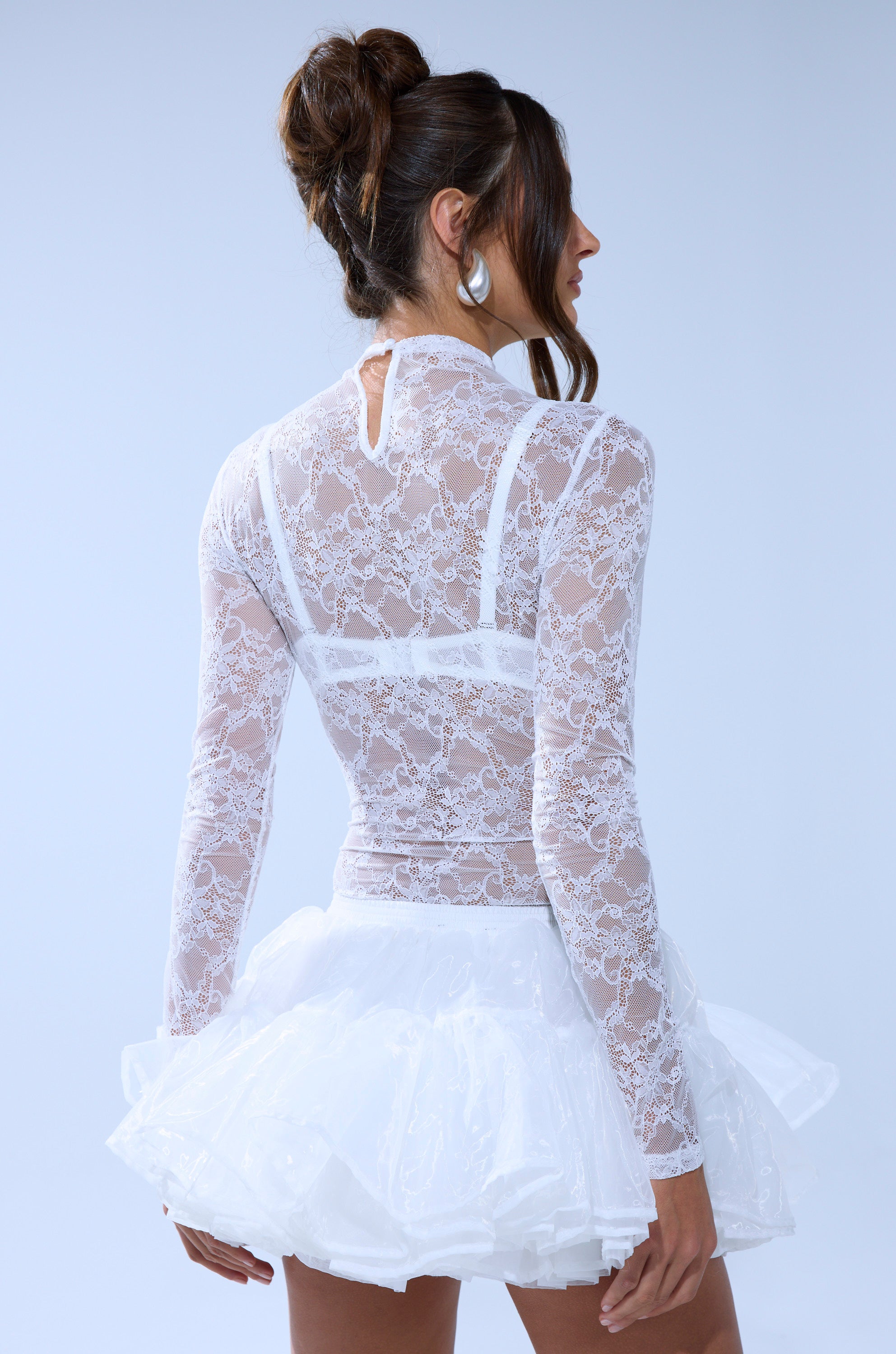 A woman with brown hair in an updo models the EVERYDAY MOCK NECK LACE BODYSUIT IN WHITE, featuring long sleeves and sheer lace, paired with a short white ruffled skirt. She faces away, showing the back of her outfit against a light blue background. 4