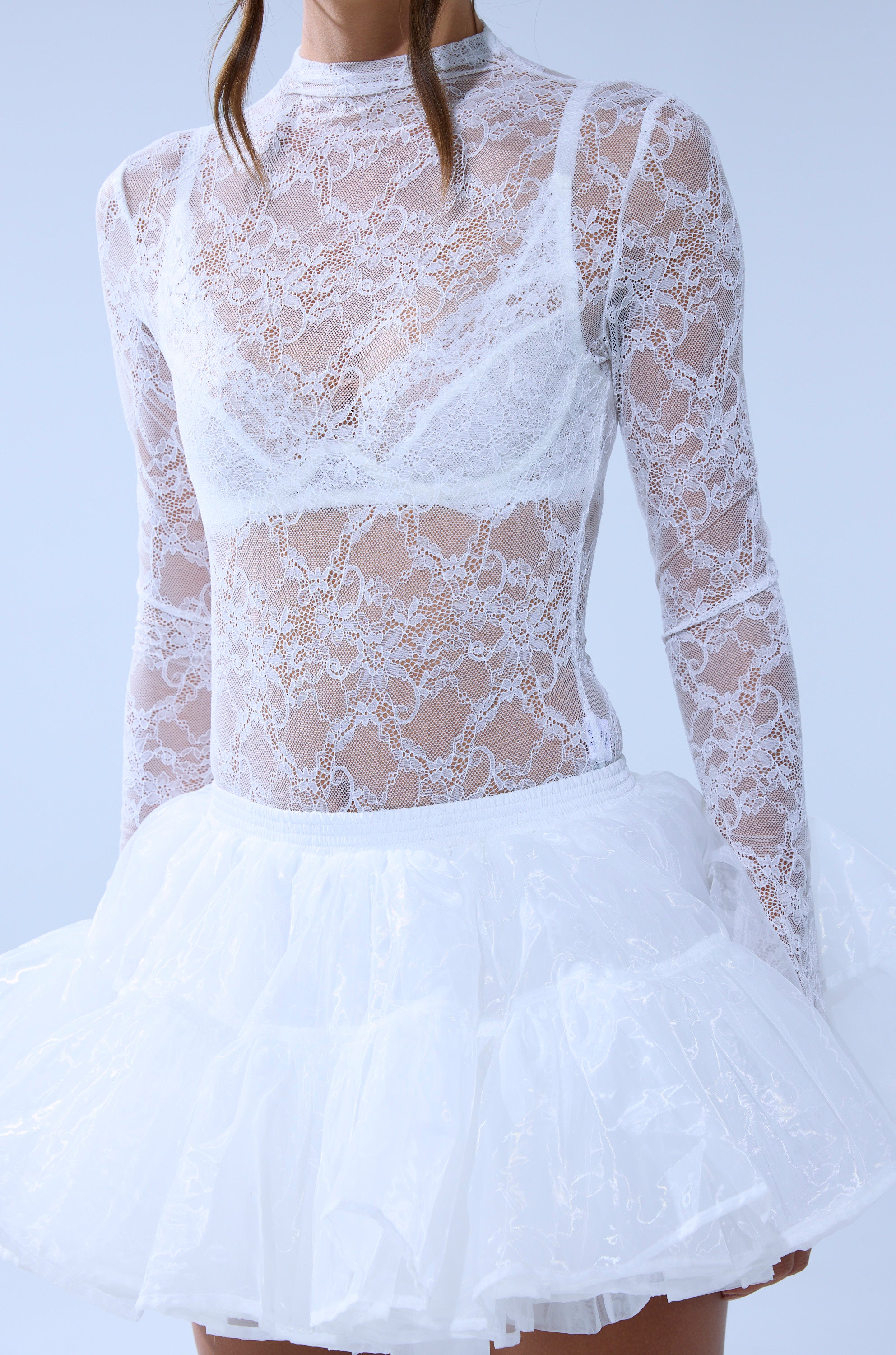 A person wears the EVERYDAY MOCK NECK LACE BODYSUIT IN WHITE with long sleeves over a white bra, paired with a ruffled, semi-transparent skirt. The background is light blue, and their face is partially out of frame. 8