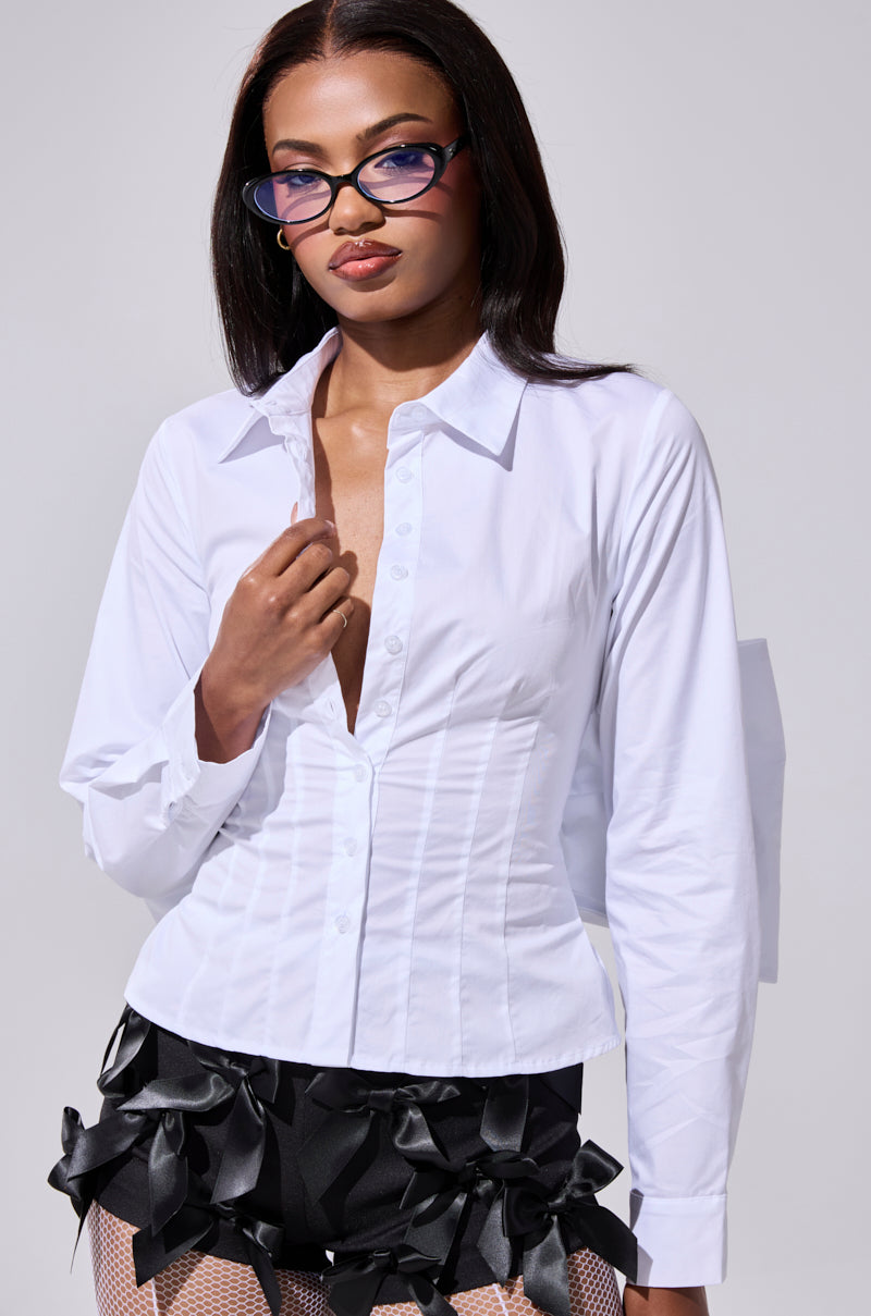 A woman poses confidently in the STRAIGHT TO THE POINT BUTTON DOWN, styled with cat-eye glasses and black ribbon-detailed shorts, against a light gray background—showcasing the blouse's chic versatility.