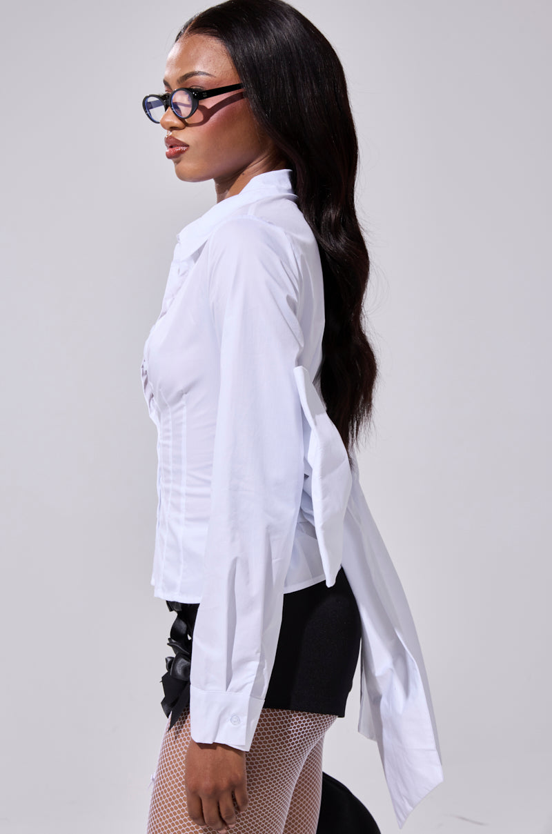 A woman with long dark hair and glasses poses in profile against a plain gray background, wearing the STRAIGHT TO THE POINT BUTTON DOWN blouse, black shorts, and fishnet stockings.
