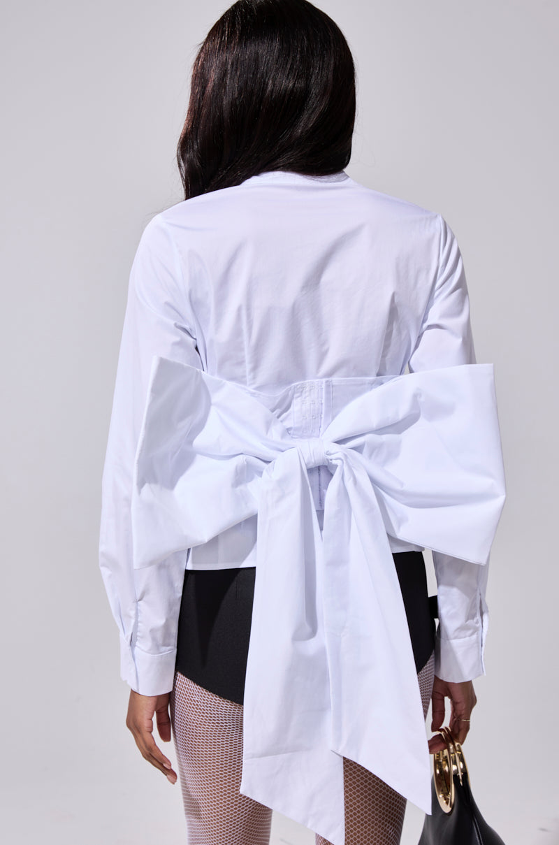 A person with long dark hair, seen from behind, wears the STRAIGHT TO THE POINT BUTTON DOWN blouse adorned with an oversized back bow, black bottoms, and carries a small gold-handled bag.