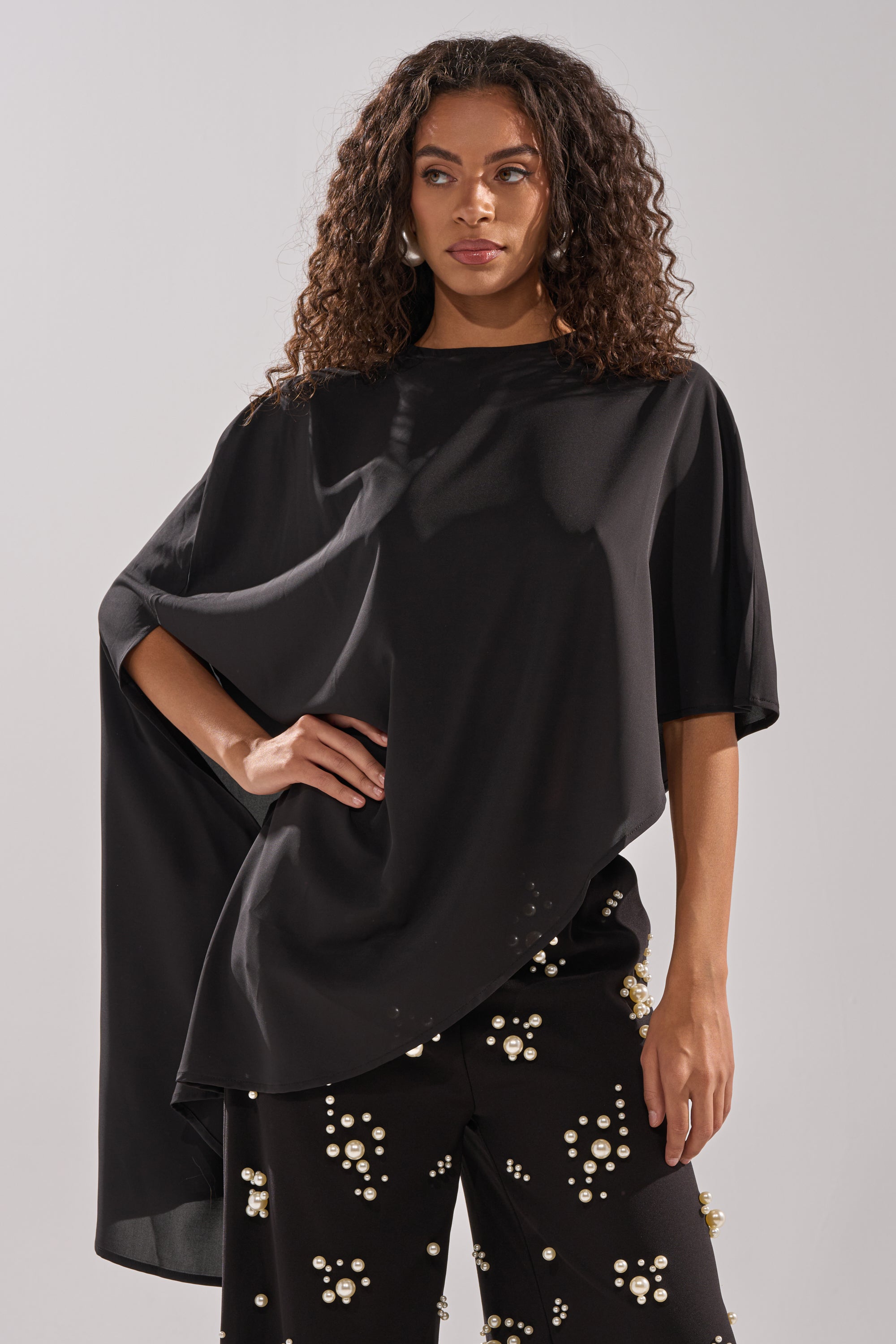 A woman with curly hair models the SOMETHING DIFFERENT OVERSIZED ASYMMETRICAL TEE and black pearl-embellished pants, posing with one hand on her hip against a plain background.
