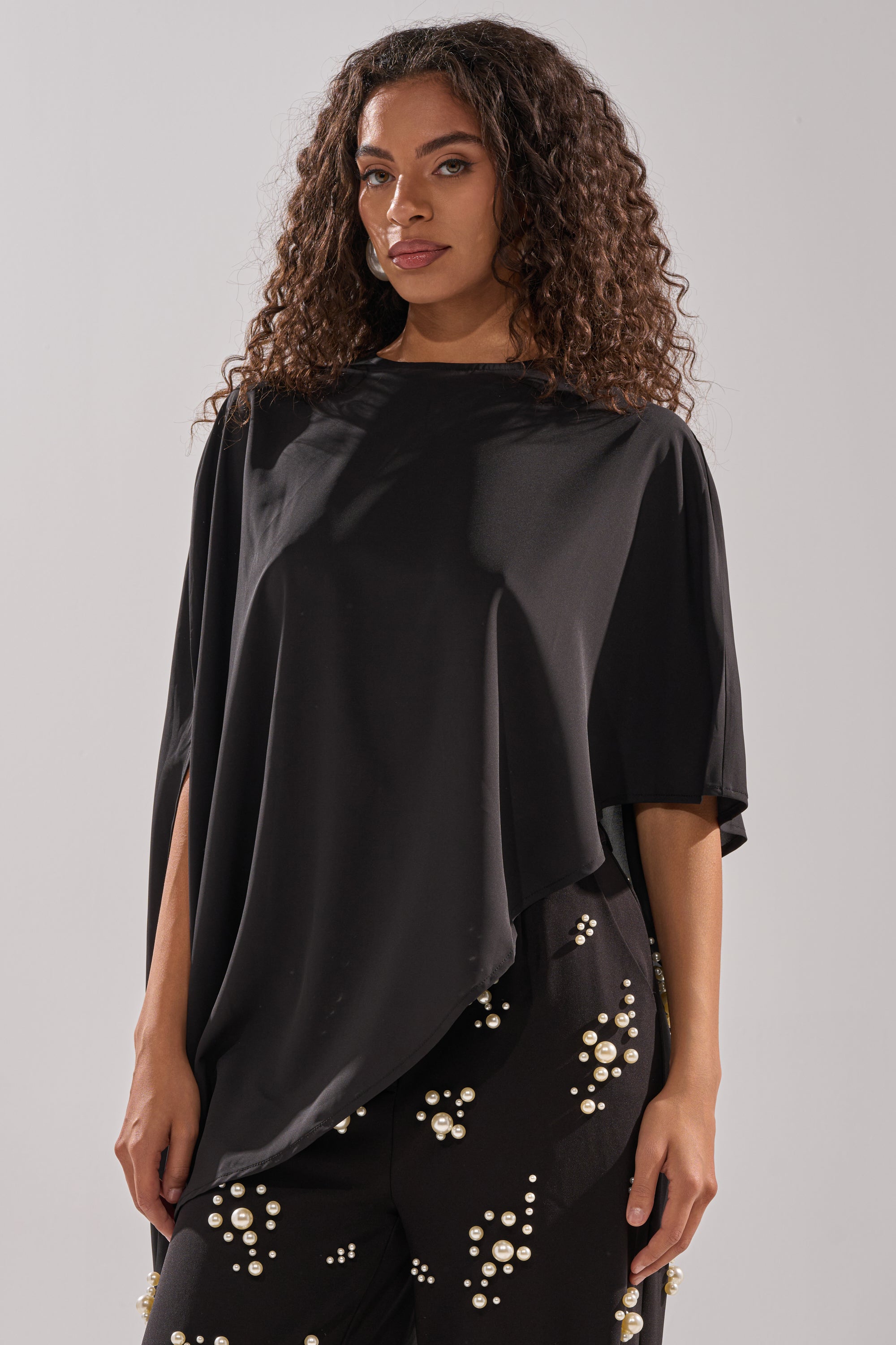 A woman with long curly hair models the SOMETHING DIFFERENT OVERSIZED ASYMMETRICAL TEE and black pants decorated with white pearl clusters, standing against a plain light gray background.