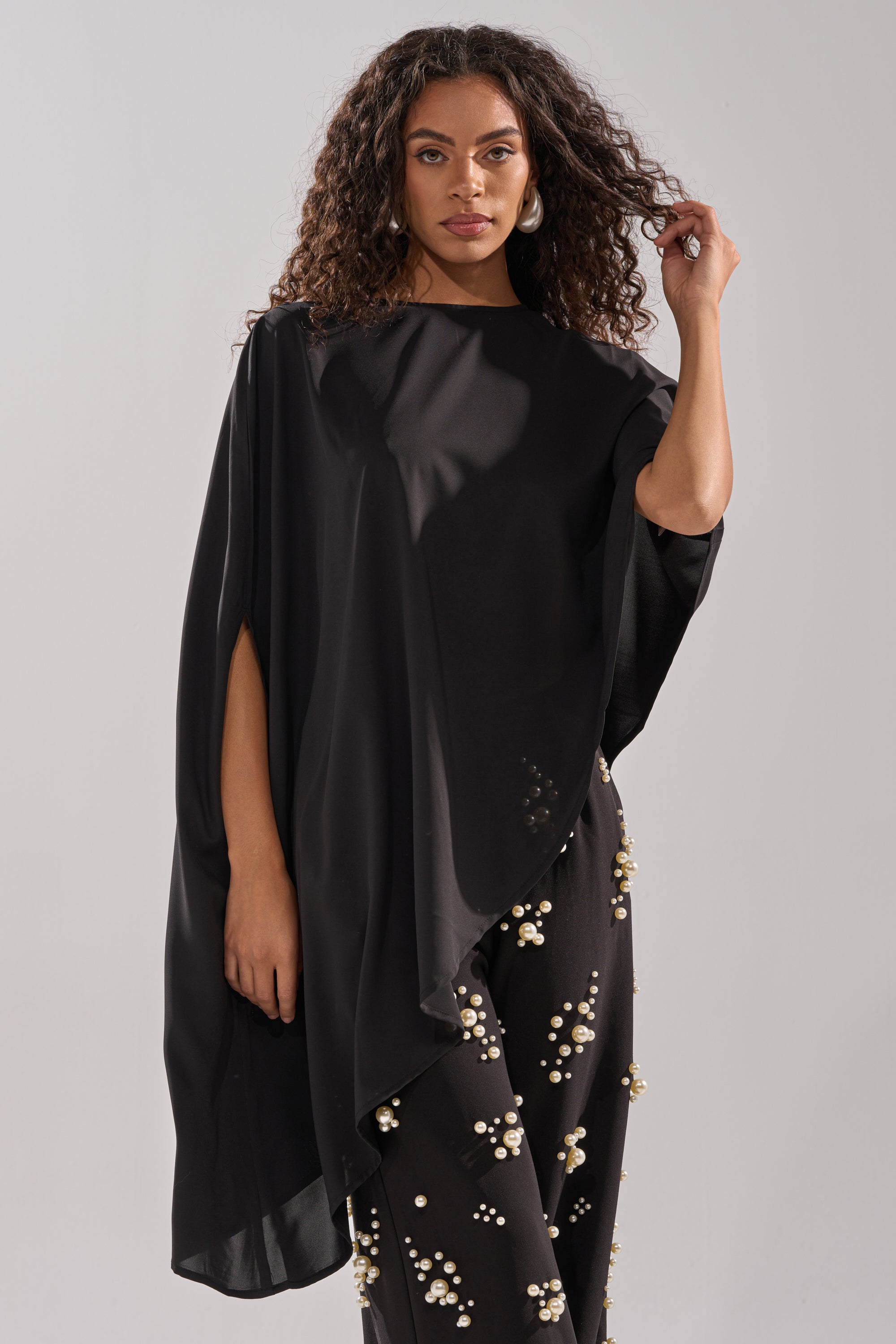 A woman with curly hair wears the SOMETHING DIFFERENT OVERSIZED ASYMMETRICAL TEE in black and black pants with white and gold bead clusters, standing against a plain light background.