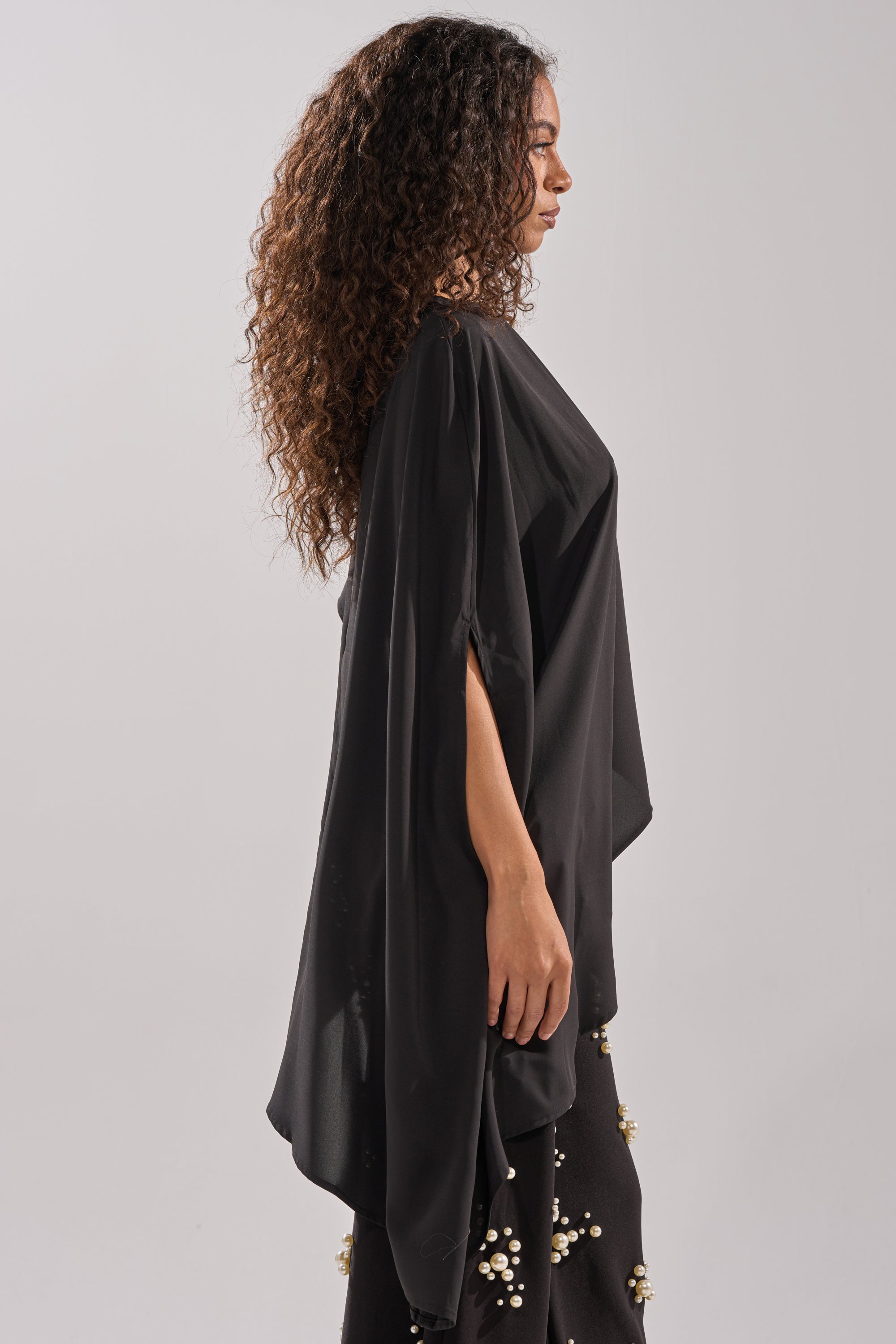 A woman with long curly hair stands in profile, wearing the SOMETHING DIFFERENT OVERSIZED ASYMMETRICAL TEE with wide sleeves and black pearl-embellished pants, against a plain light background.