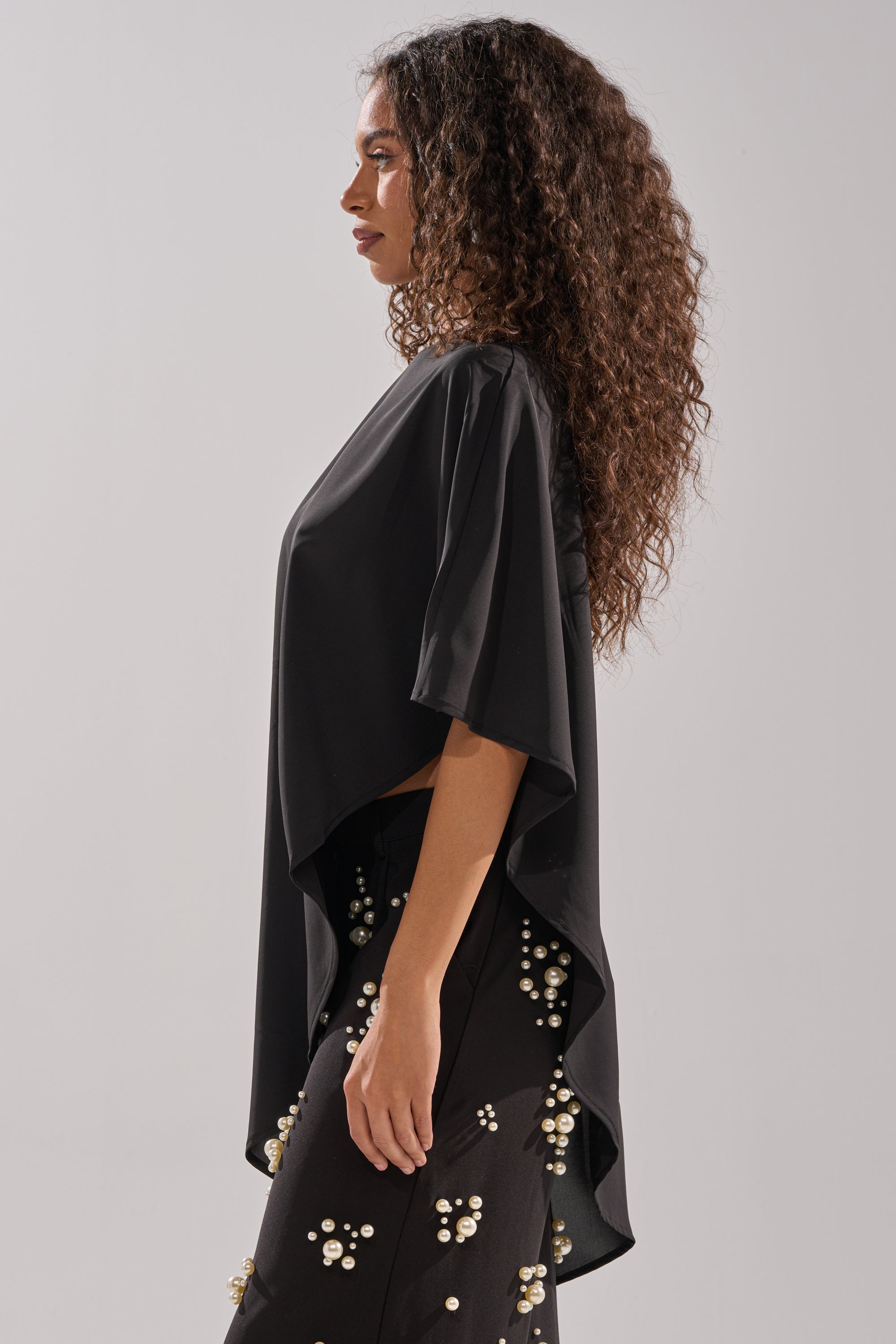 A woman with curly hair stands in profile, wearing the SOMETHING DIFFERENT OVERSIZED ASYMMETRICAL TEE with flowing cape-like sleeves and black pants featuring white floral embroidery against a plain light gray background.